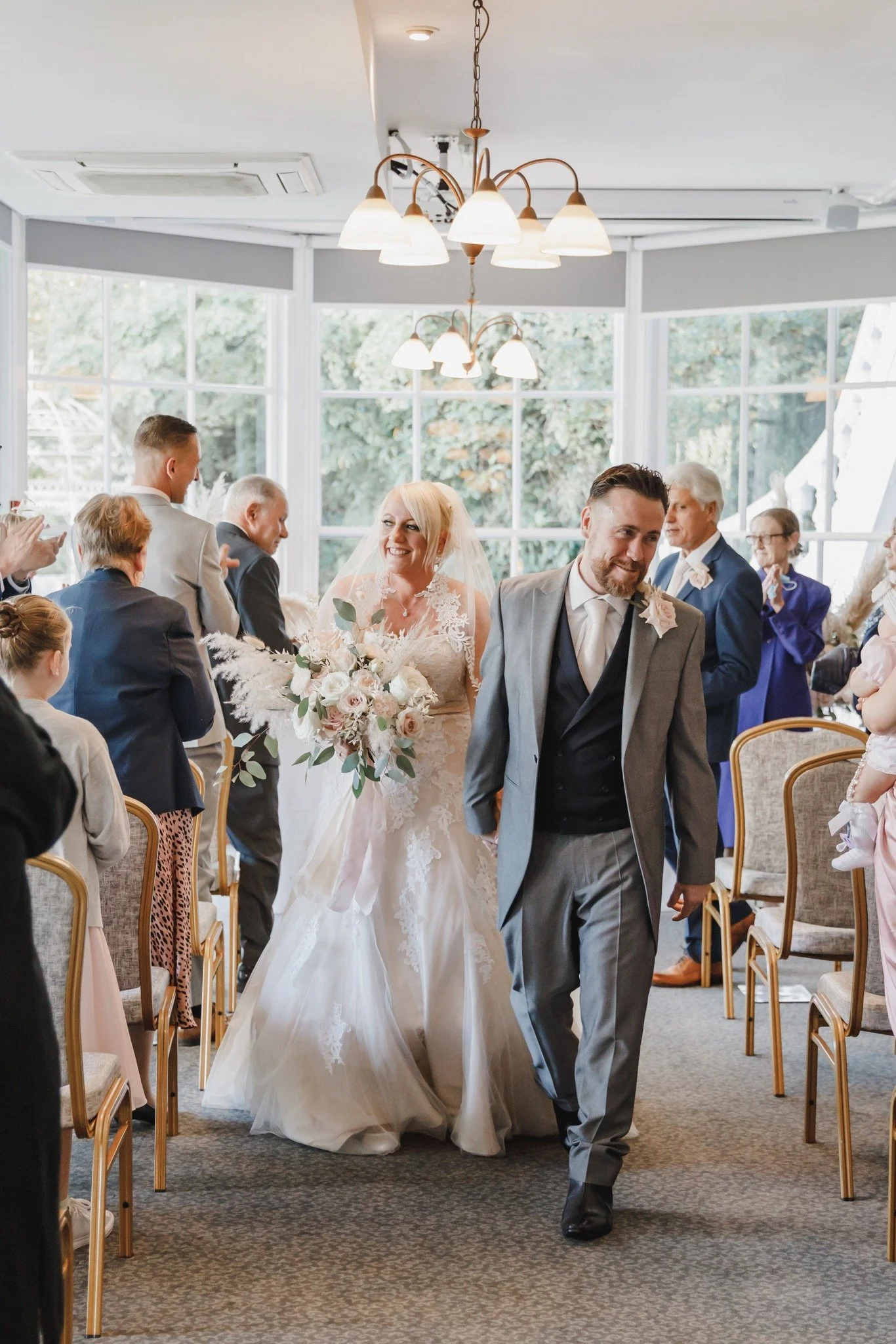 A bride and groom walking down the aisle at their wedding reception, surrounded by guests in a decorated indoor venue with large windows.