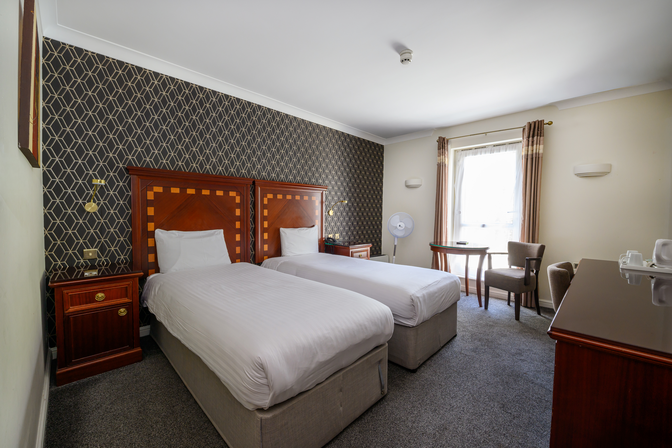 Hotel room with two beds, dark patterned accent wall, wooden headboards, nightstand, fan, chairs, and window with curtains.