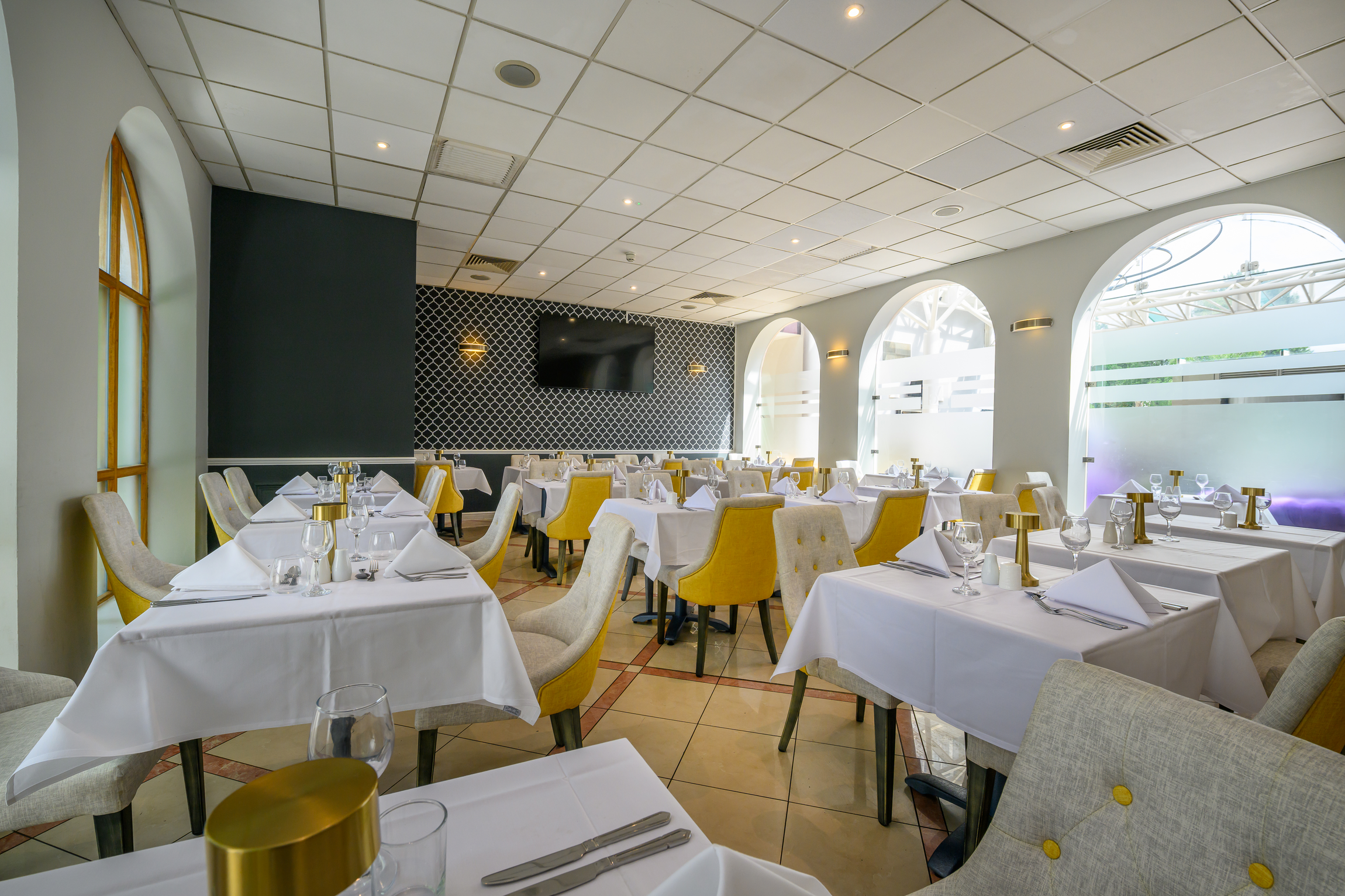 Interior of a restaurant with white tablecloths, neatly folded napkins, wine glasses, and gold-colored candle holders on each table. Yellow and beige upholstered chairs surround the tables. The ceiling has a grid pattern with embedded lighting. Large arched windows on the right side let in natural light. A television is mounted on a patterned black wall at the back of the dining area.