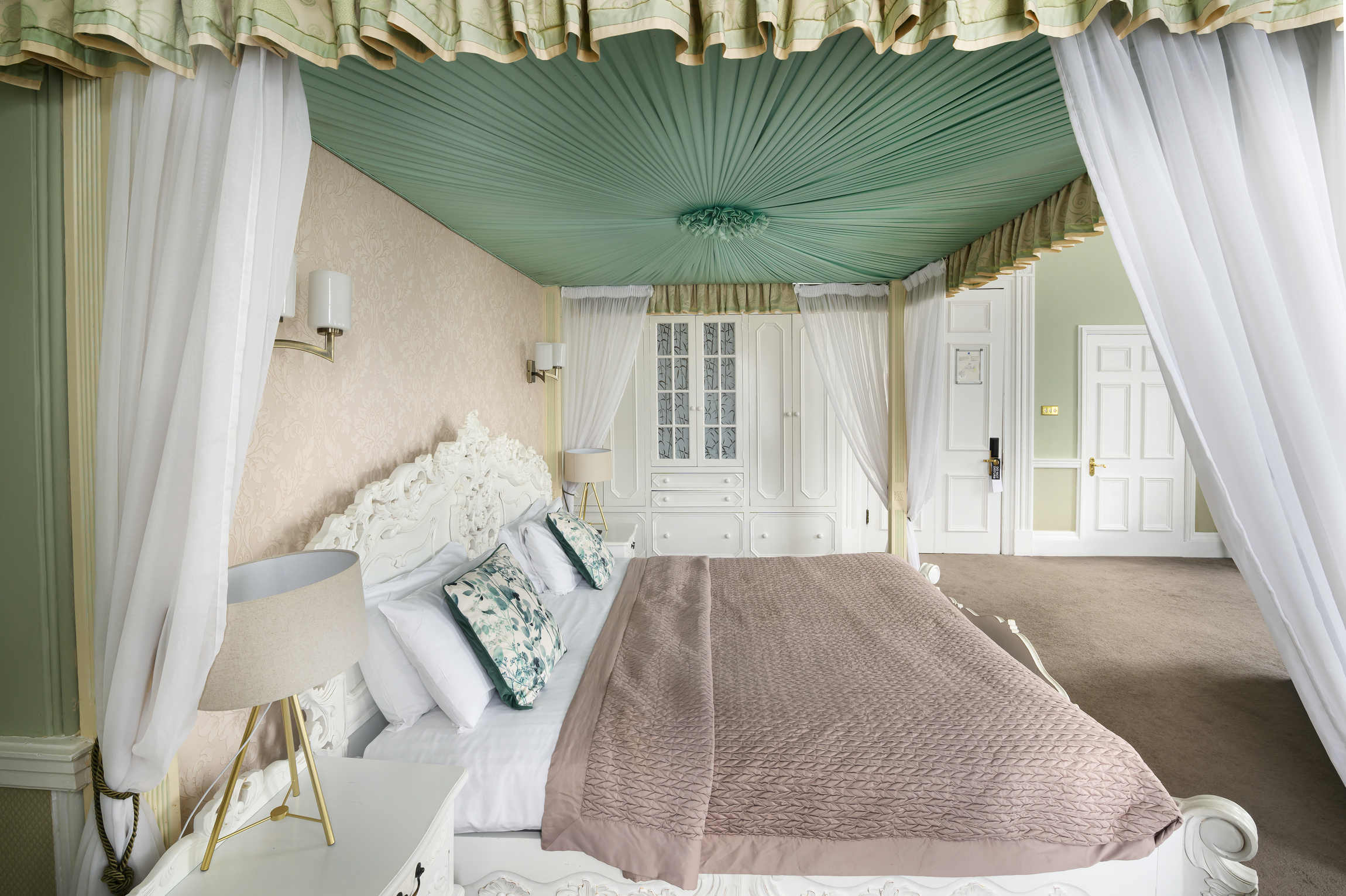 A luxurious bedroom with a four-poster bed, ornate white headboard, and green draped ceiling. The bed has multiple pillows and a brown quilt. There are bedside tables with lamps, and a large corner wardrobe with glass doors. The room features light-c