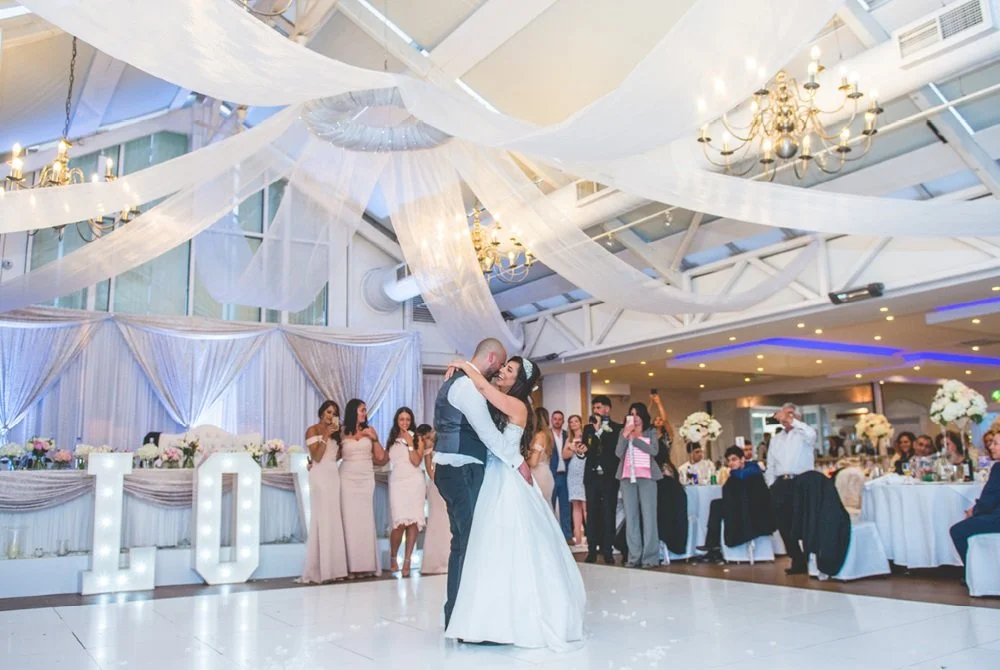 Couple dancing at a wedding reception with bridesmaids and guests watching in a decorated banquet hall.