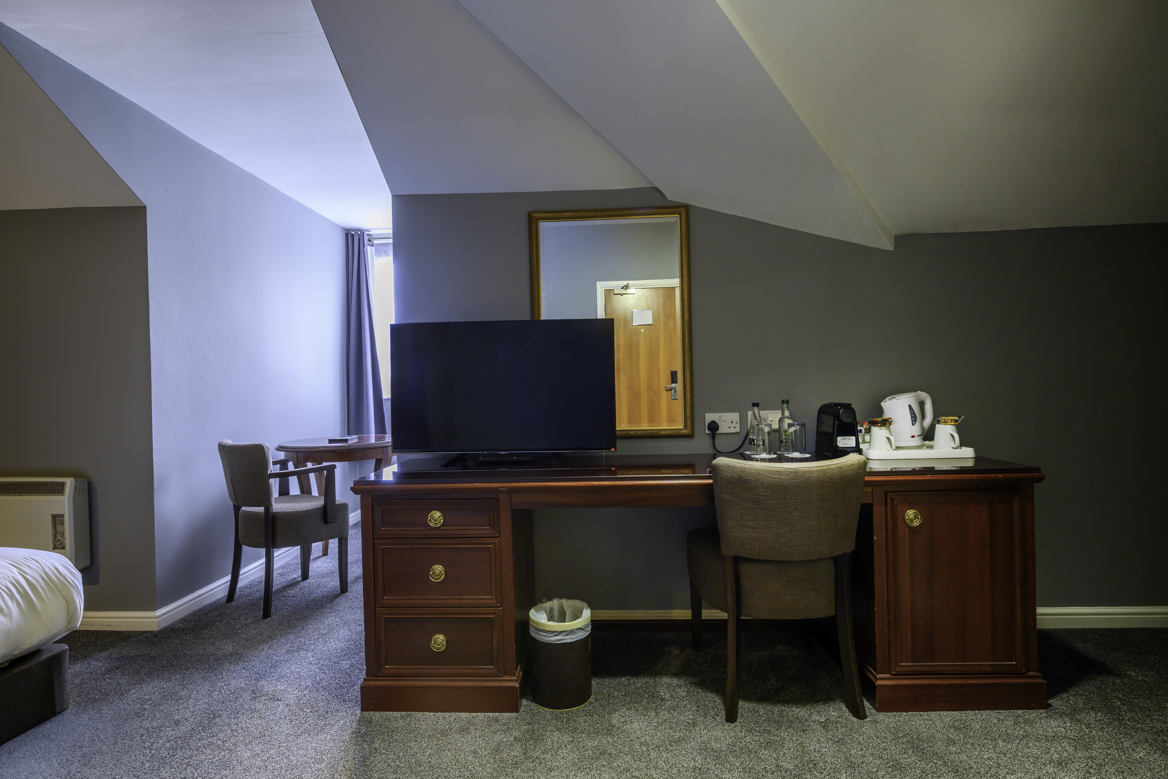 Hotel room with a wooden desk, TV, chairs, mirror, kitchenette appliances, and a bed partially visible on the left.