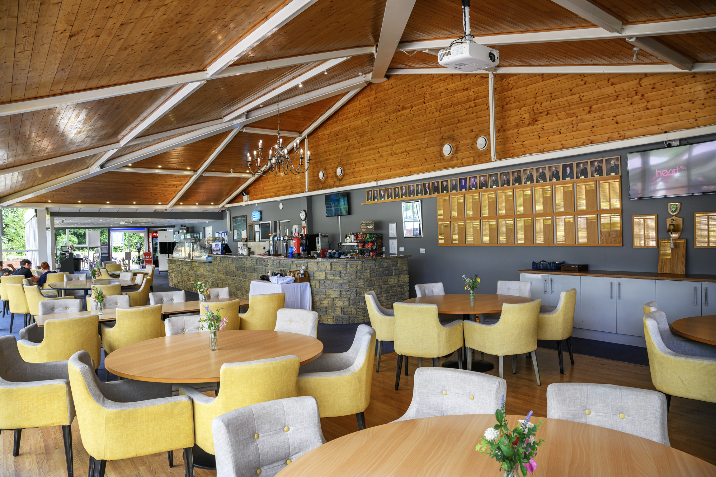 Interior of a restaurant or cafe with yellow and gray upholstered chairs around wooden tables, a stone bar counter, and a wood-paneled vaulted ceiling with a chandelier. Decorations include framed photographs on the wall and flowers on the tables.