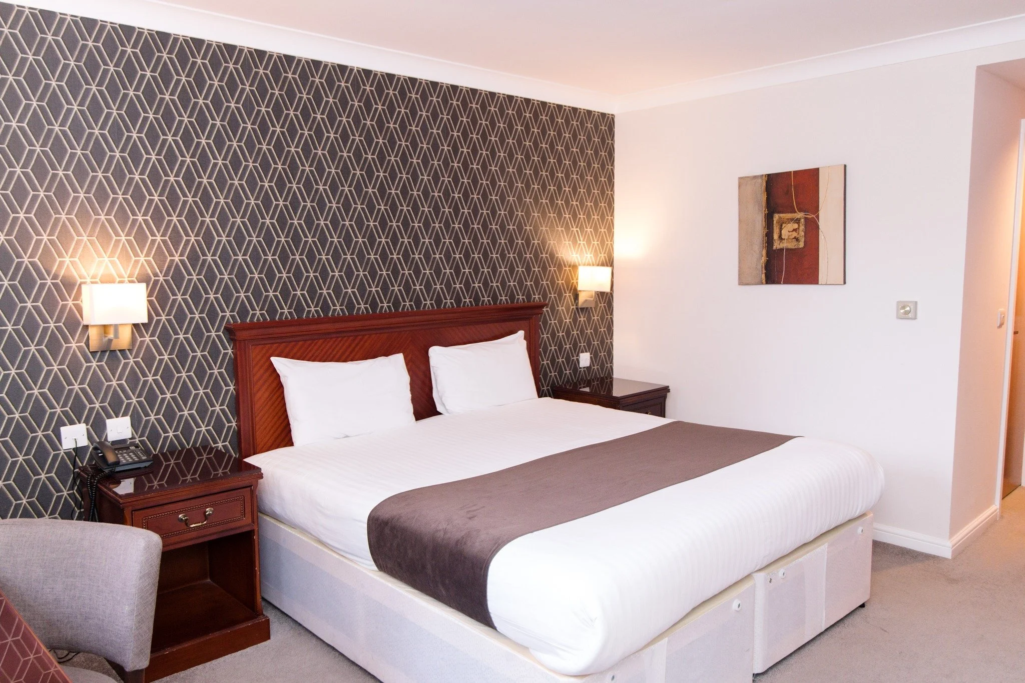 Hotel room with a large bed, dark patterned accent wall, white bedding, and wooden headboard, two bedside tables with lamps, wall art, and beige carpet.