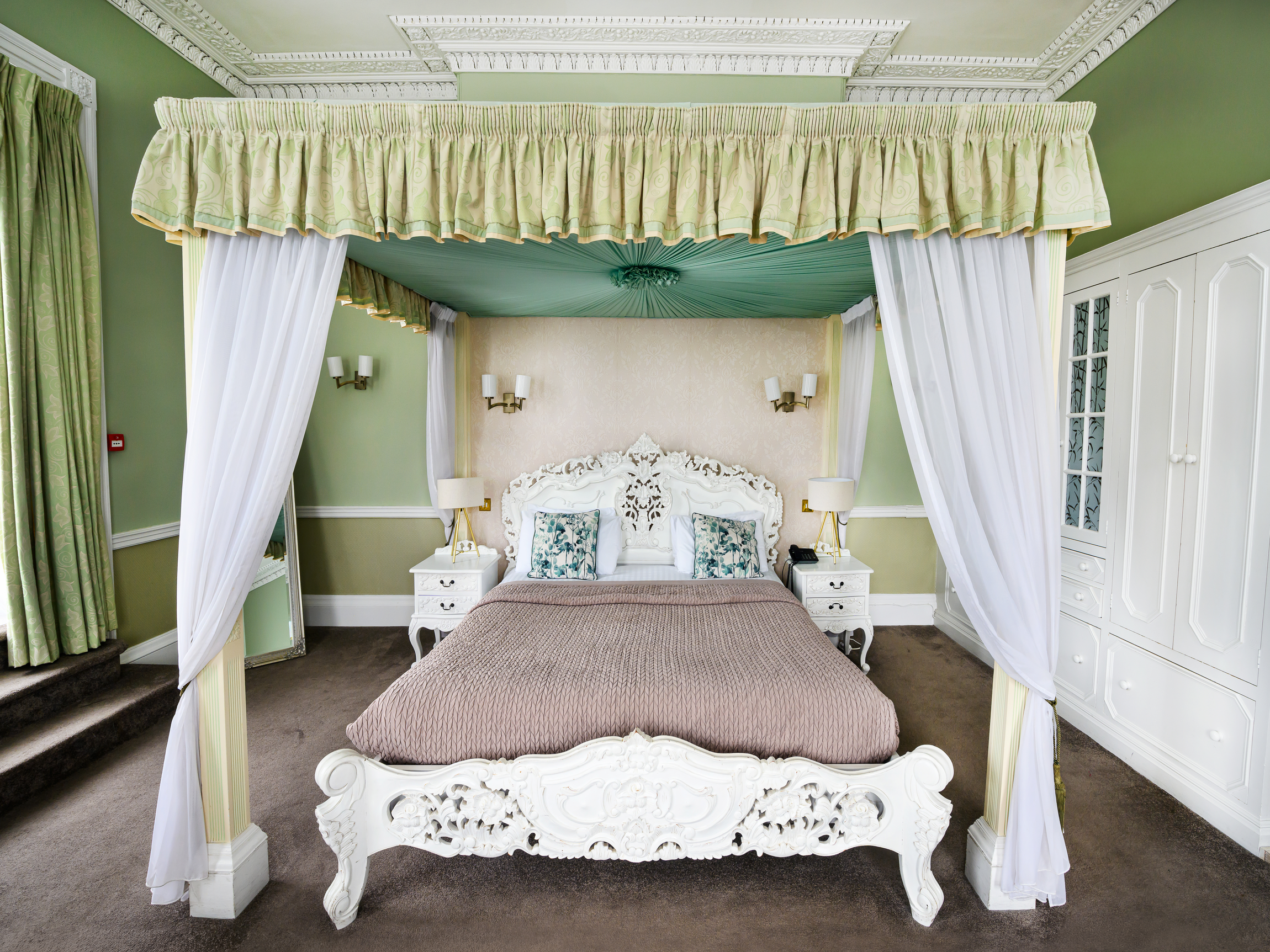A luxurious bedroom with a white ornate four-poster bed, cream-colored headboard, and light bedding. The room features green walls, decorative crown molding, and a canopy with green fabric and white curtains. There are matching white bedside tables w