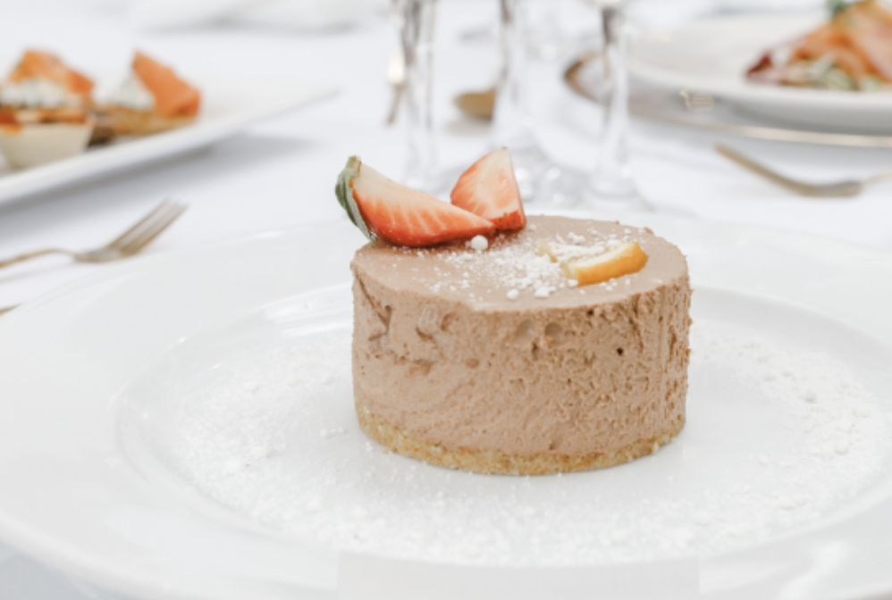 A small round strawberry mousse dessert garnished with a sliced strawberry on top, served on a white plate with a light dusting of powdered sugar, set on a white table with wine glasses and plates in the background.