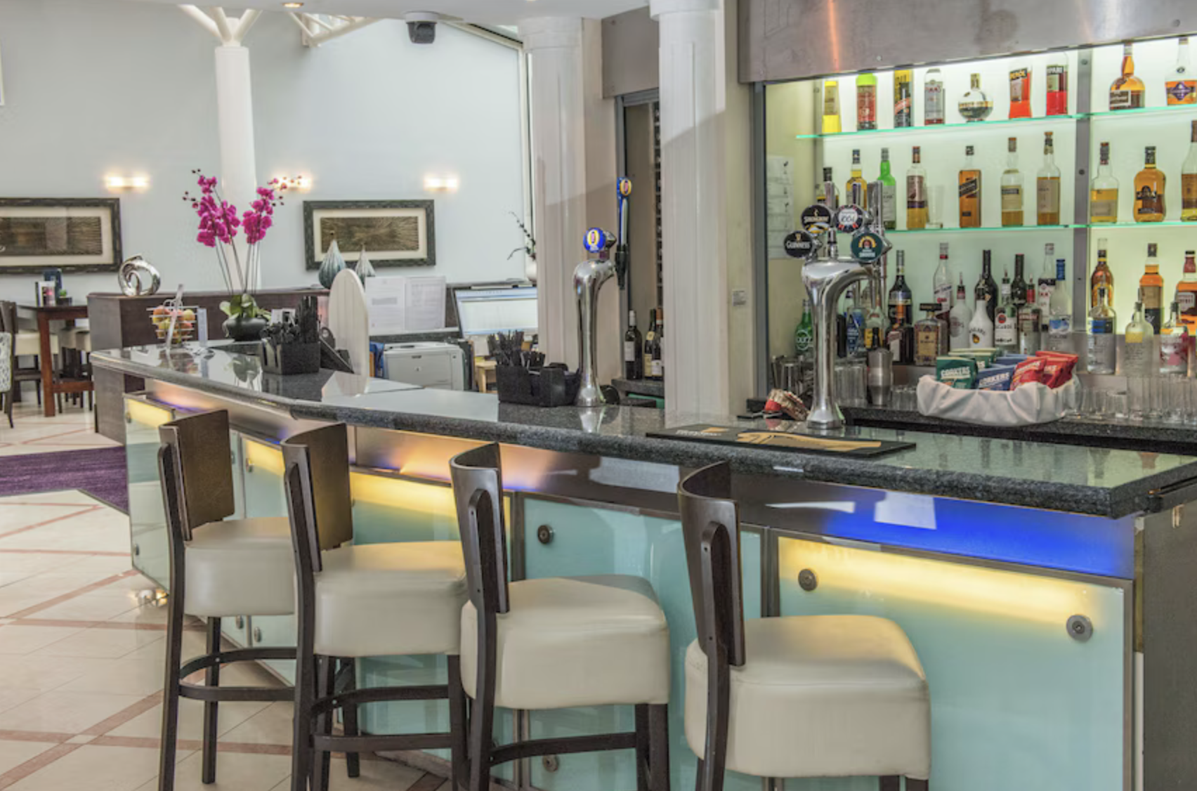 Empty bar counter with chairs, liquor bottles on shelves, and a basket of chips.