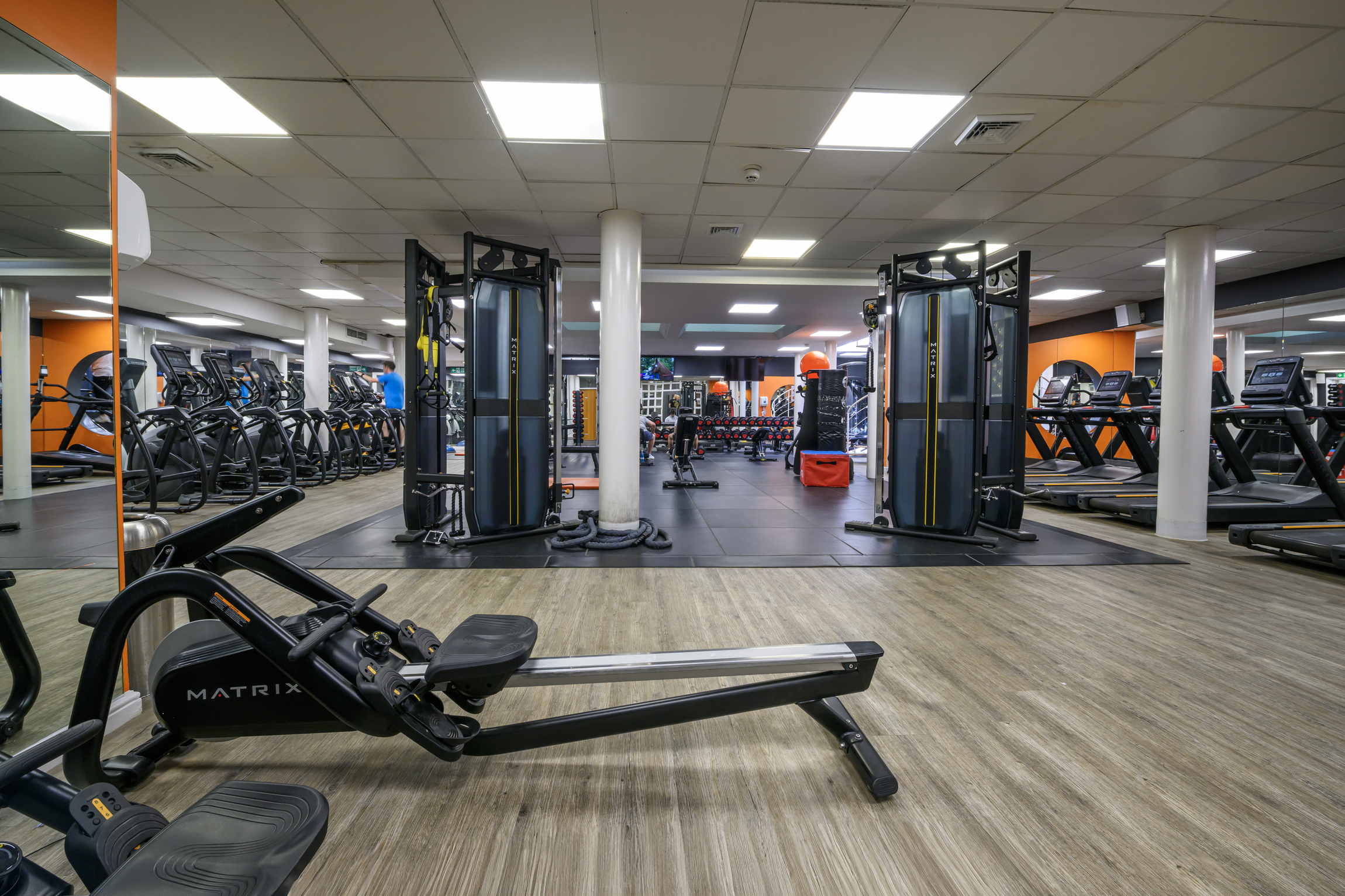 Empty gym with various exercise equipment including rowing machine in the foreground, treadmills and ellipticals along the sides, and strength training gear in the back, illuminated by ceiling lights.