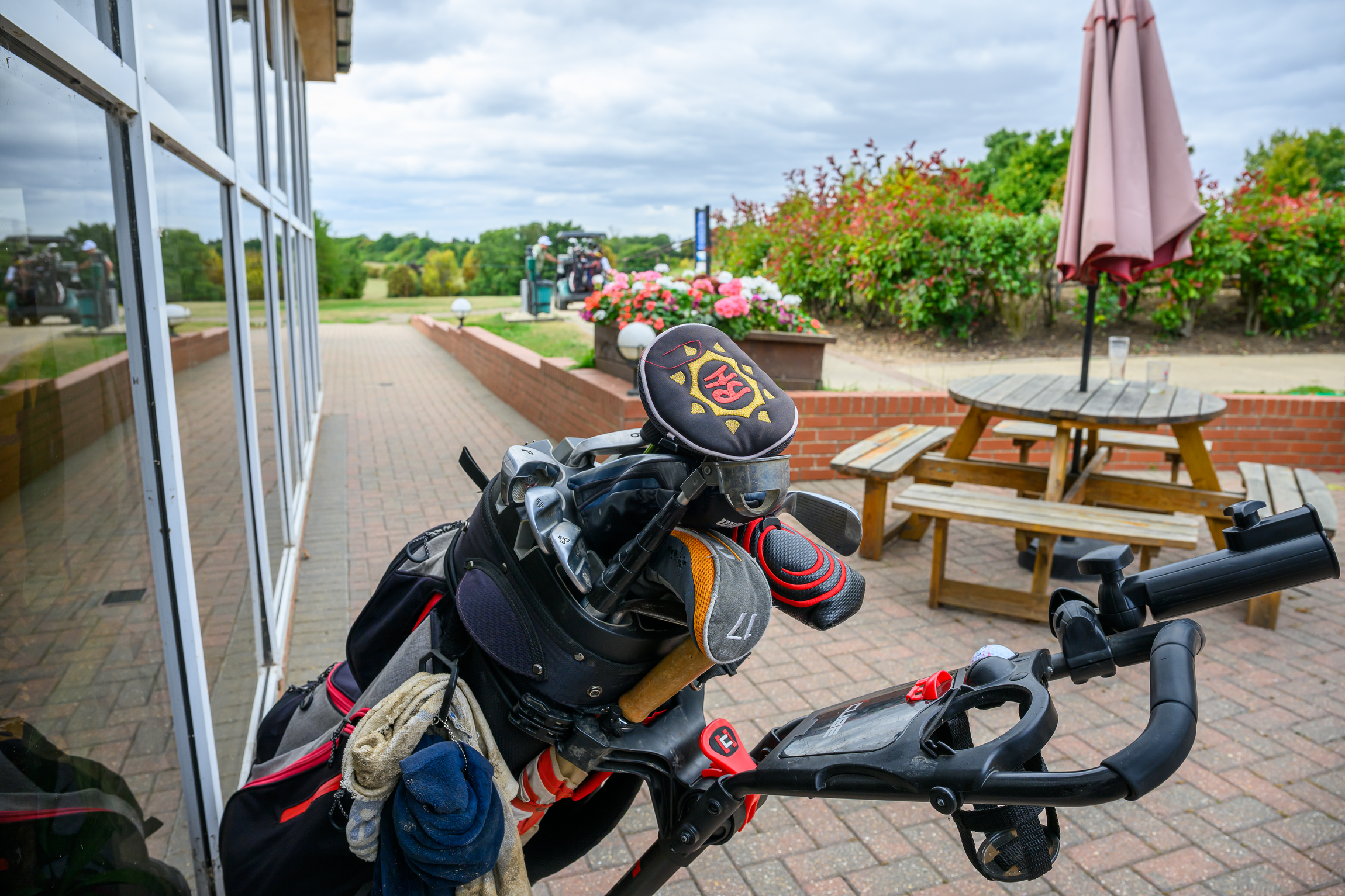 Golf bag with clubs and accessories placed on a golf push cart outside near a patio area with picnic tables, umbrella, flowers, and golf course in the background.
