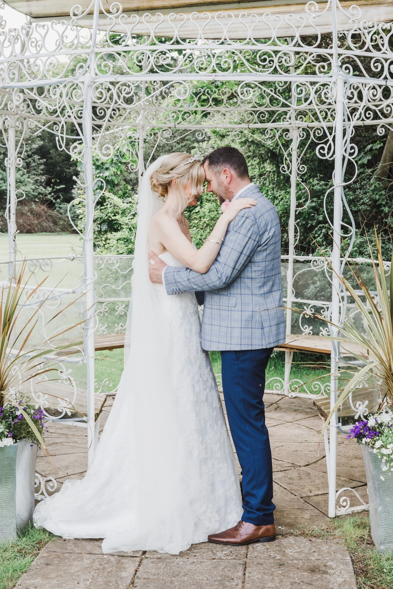 A bride and groom embrace under a white metal gazebo in a garden, with lush greenery and potted flowers nearby.