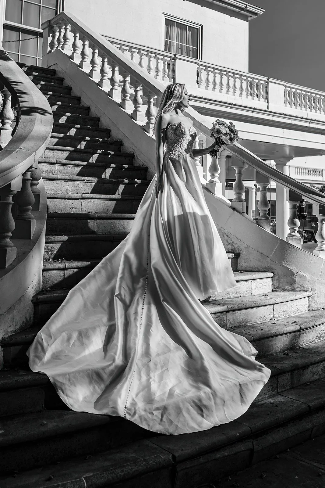 A bride wearing a wedding dress with a long train, holding a bouquet, ascending outdoor staircase with railing.