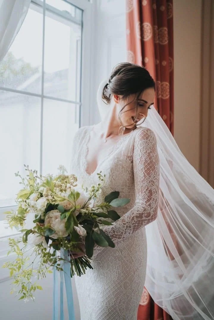 A bride with dark hair styled in an updo, wearing a long-sleeved lace wedding gown, holding a large bouquet of white and green flowers, standing next to a window with natural light, and a pink curtain with a gold pattern hanging behind her.