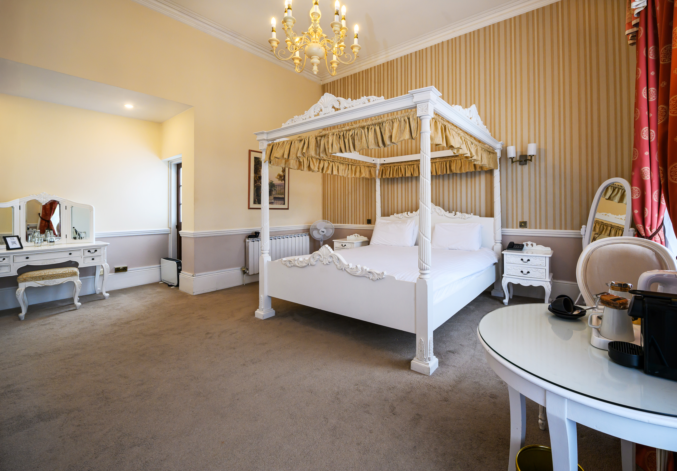 A luxurious hotel room featuring a white ornate four-poster bed with golden canopy and striped wallpaper, a chandelier, a dressing table with mirror, a side table with a fan, and a round table with coffee-making equipment.