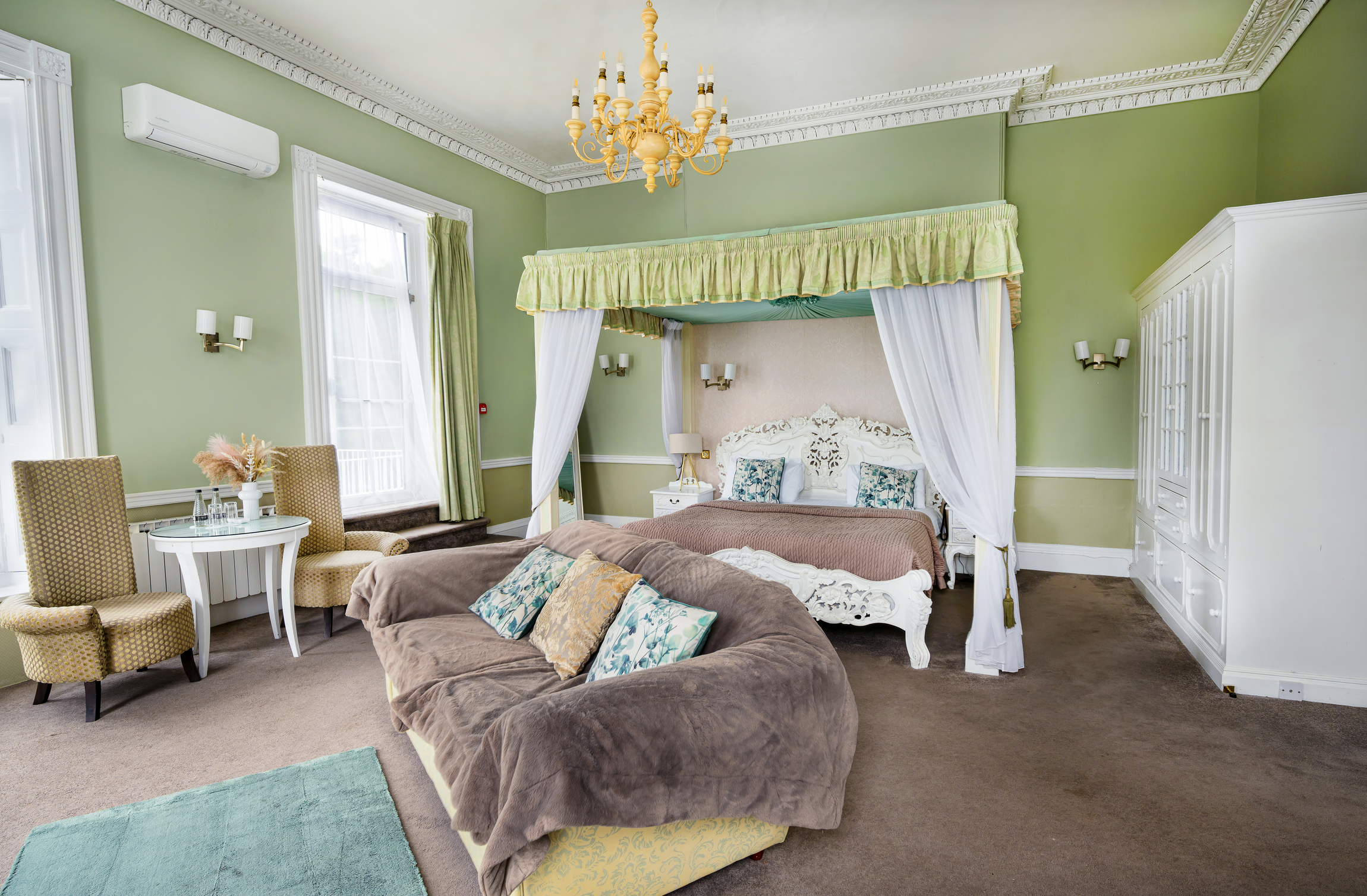 Bright hotel room with green walls, white crown molding, large windows, a chandelier, a canopy bed with white curtains, a brown sofa with pillows, a small white table with chairs, and a white wardrobe.