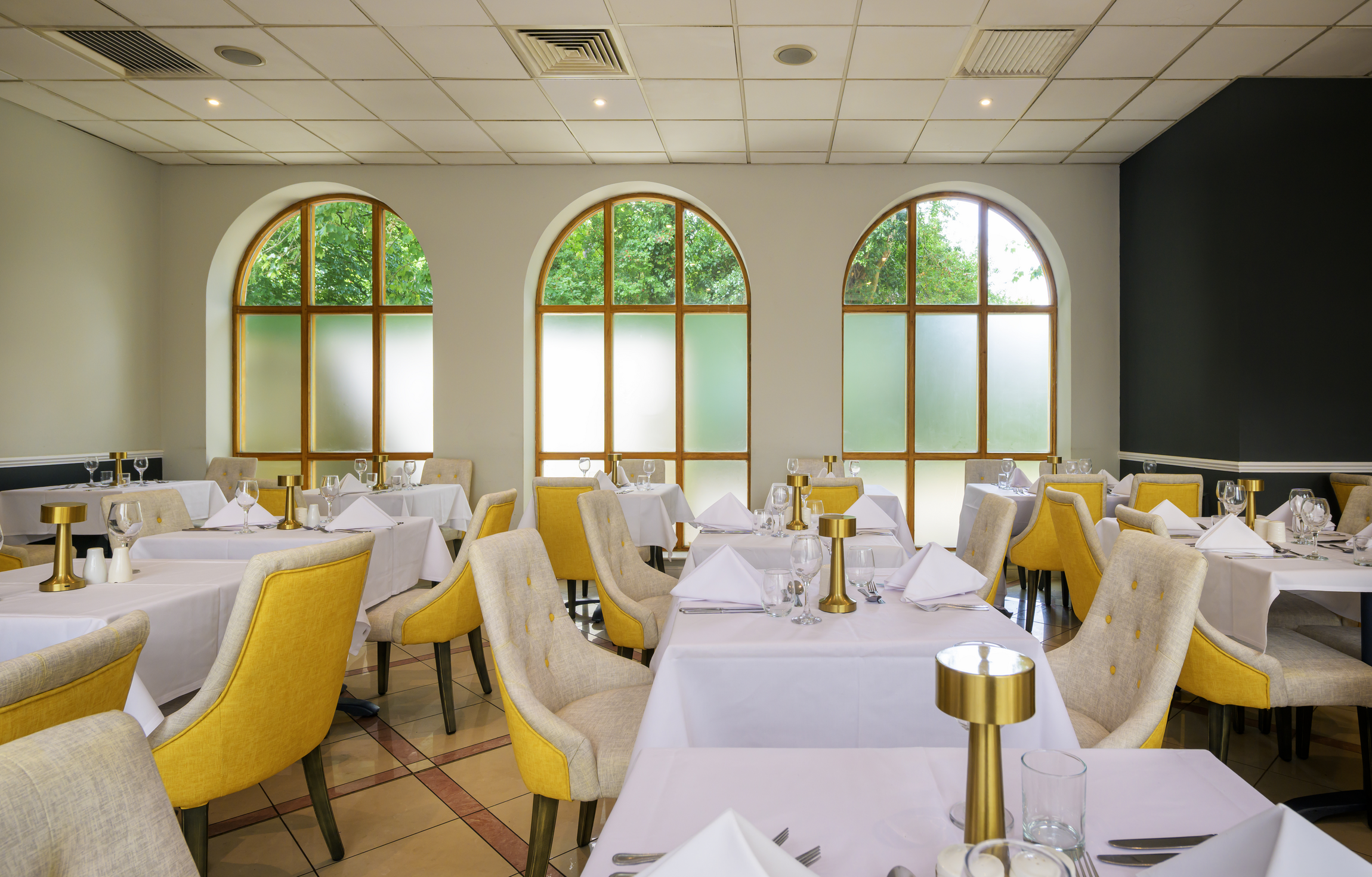 Elegant dining room with white tablecloths, yellow and beige upholstered chairs, tall arched windows with frosted glass, set for a formal meal with wine glasses, napkins, and gold candle holders.
