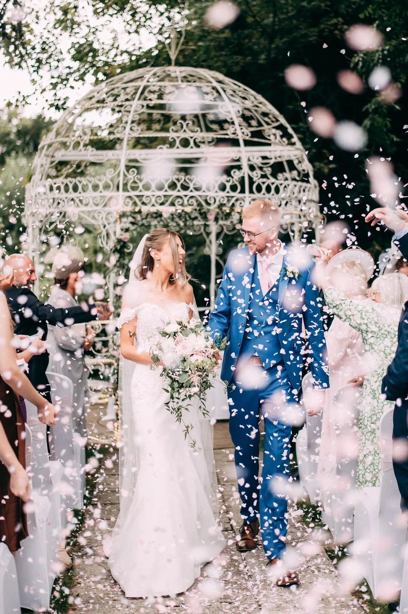 Bride and groom walking down aisle at their wedding, surrounded by guests throwing confetti, under a decorative metal gazebo, outdoors.