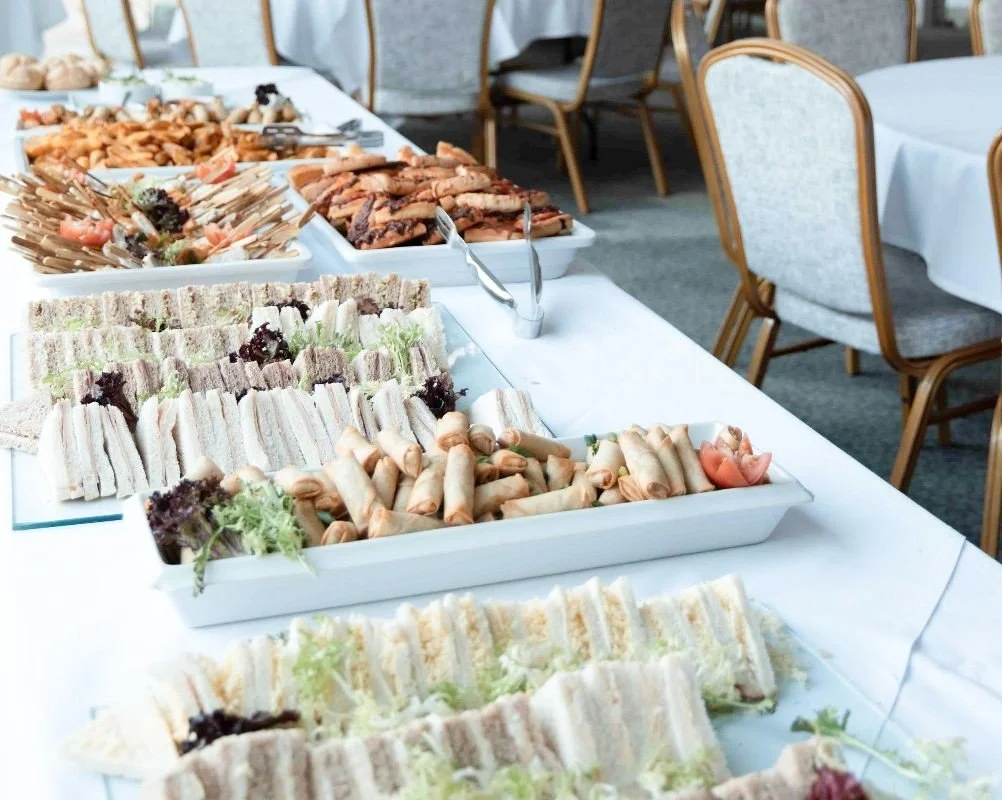 A buffet table with various finger foods and sandwiches, set in an event space with chairs and tables.