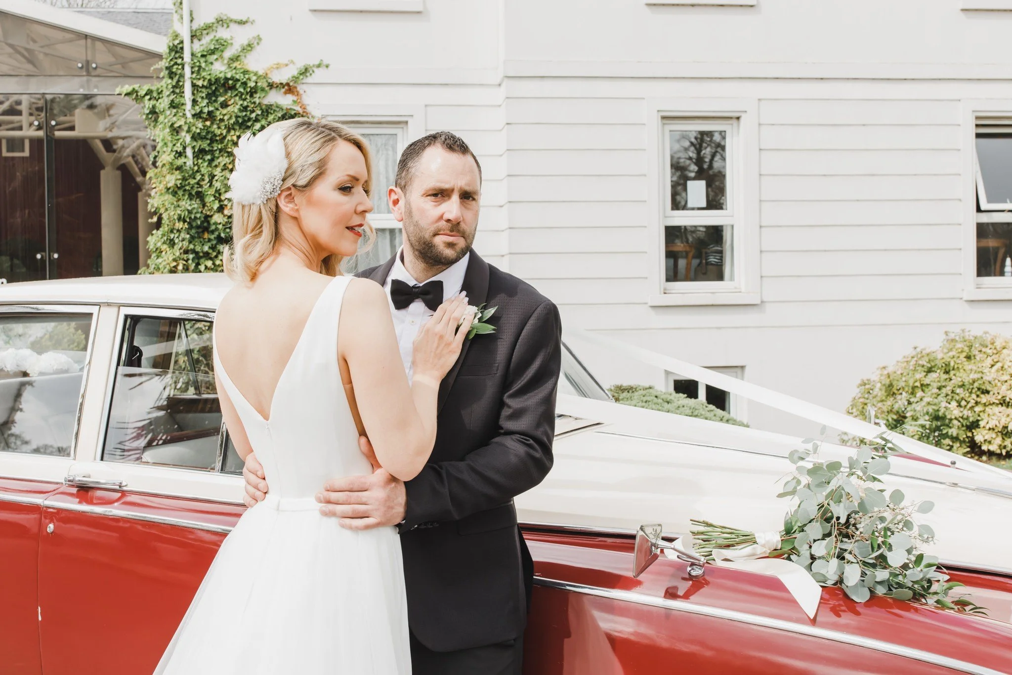 A bride and groom standing in front of a vintage car decorated with flowers outdoors, with a white wooden house in the background.