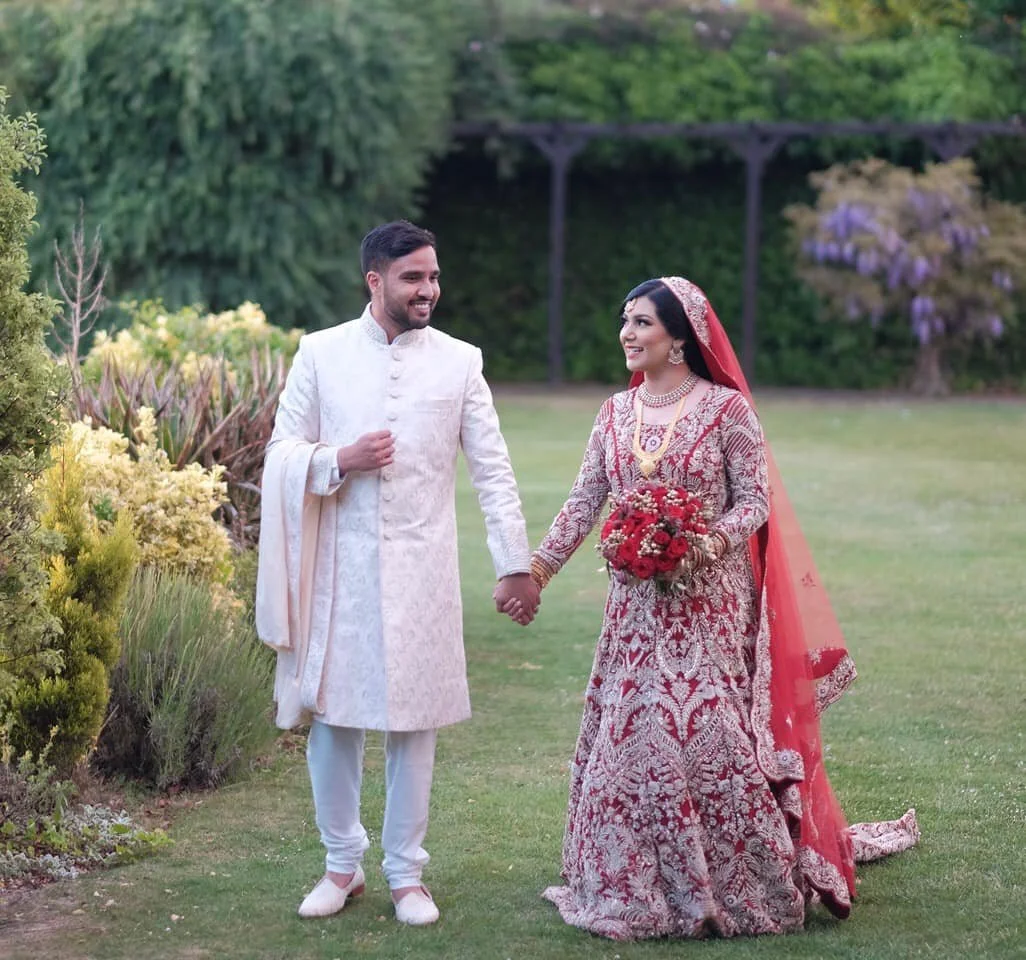 A newlywed couple dressed in traditional South Asian wedding attire walking hand in hand in a garden, smiling at each other. The bride is holding a bouquet of red flowers and wearing red and gold embroidery, while the groom is dressed in white with s