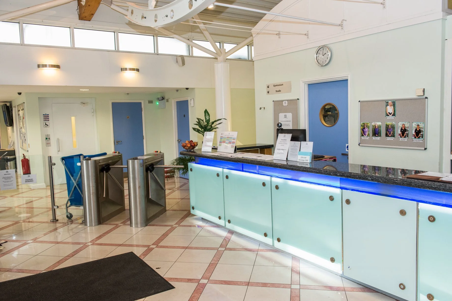 Hospital reception desk with computer, sign-in sheets, and a bowl of fruits, with security turnstiles in front, and doors, bulletin boards, and a clock on the wall in the background.