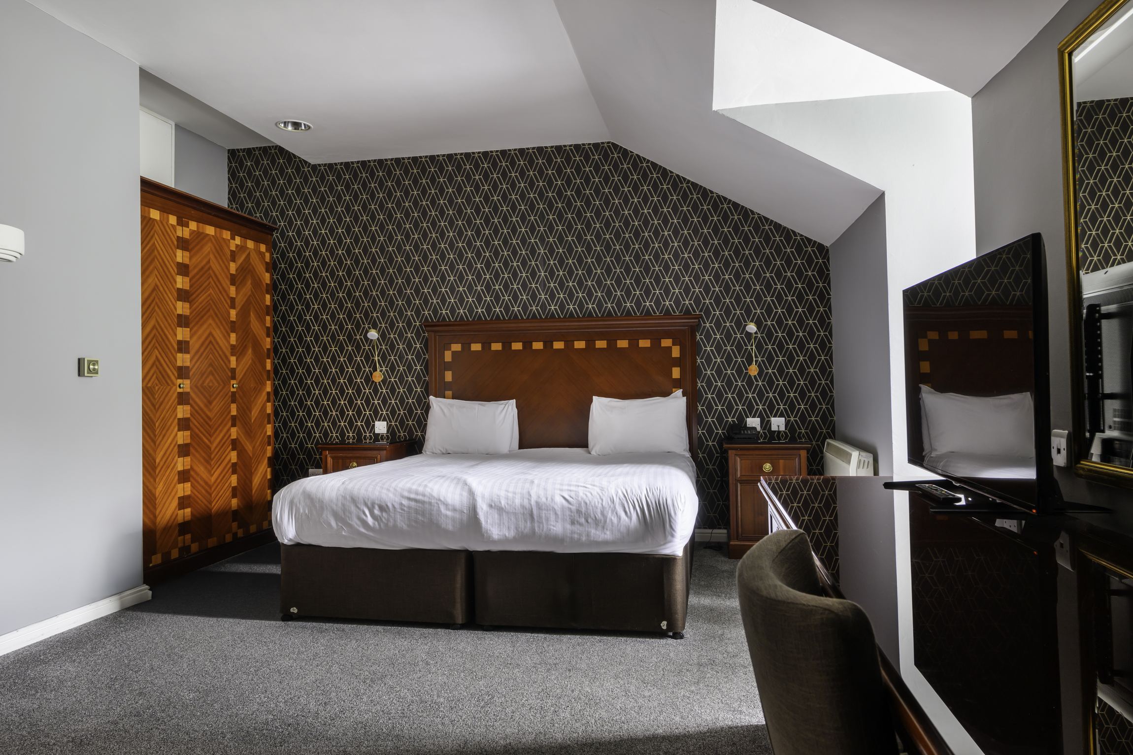 A hotel room with a double bed, wooden headboard, black and gold accent wallpaper, two bedside tables with lamps, a flat-screen TV, a chair, and an angled ceiling with a skylight.