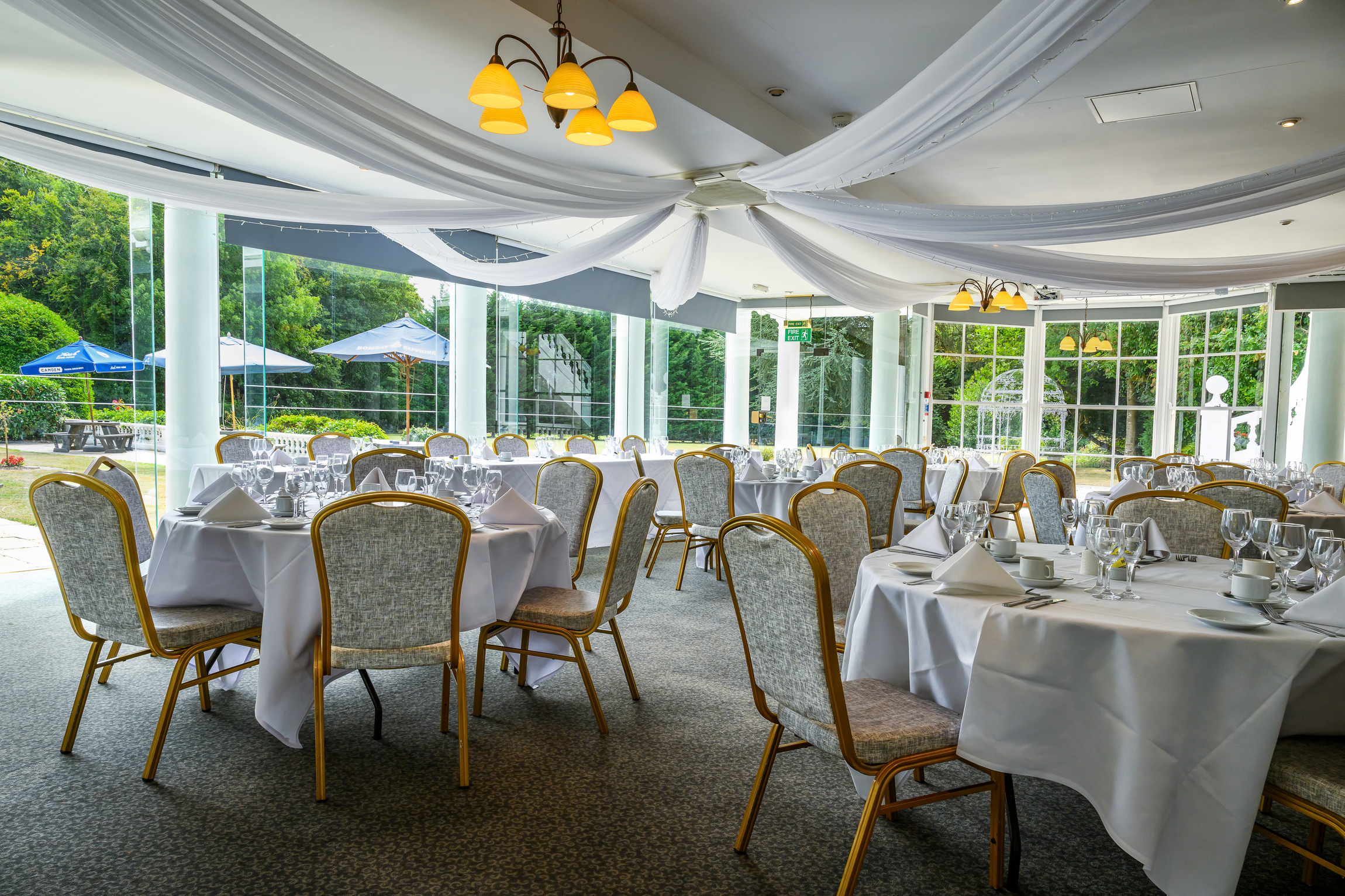 Elegant dining room set up with round tables covered in white tablecloths, each set with white plates, cups, utensils, and multiple wine glasses. Several chairs with beige upholstery and gold-colored frames surround the tables. The room has large glass windows with a view of a patio area with umbrellas and trees outside. White drapery adorns the ceiling, and warm yellow lighting fixtures hang from above.
