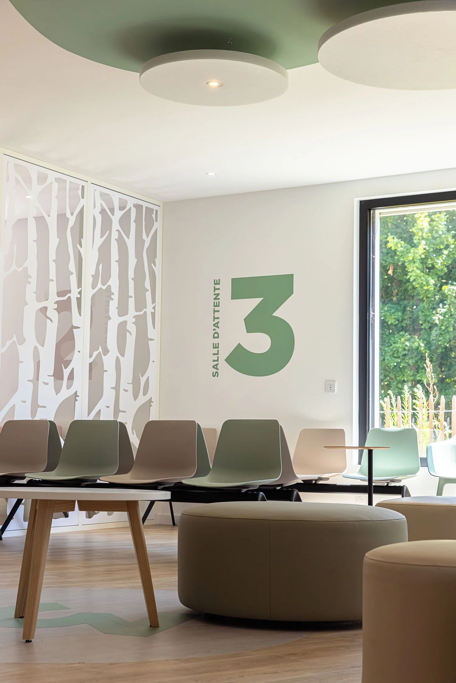 Salle d'attente du Centre Main Morbihan avec des chaises modernes, un mur décoratif en forme d'arbres, et une grande fenêtre laissant entrer la lumière naturelle.