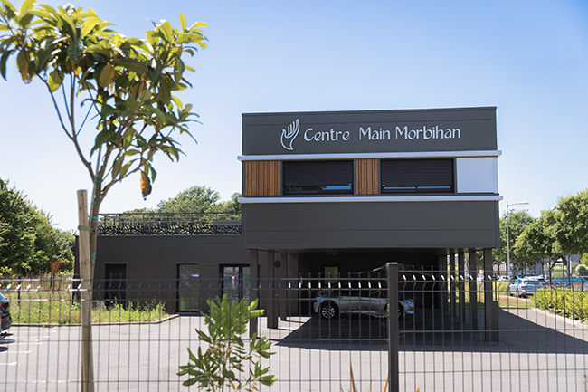 Bâtiment moderne du Centre Main Morbihan avec un parking extérieur, entouré de jeunes arbres et de végétation sous un ciel clair.
