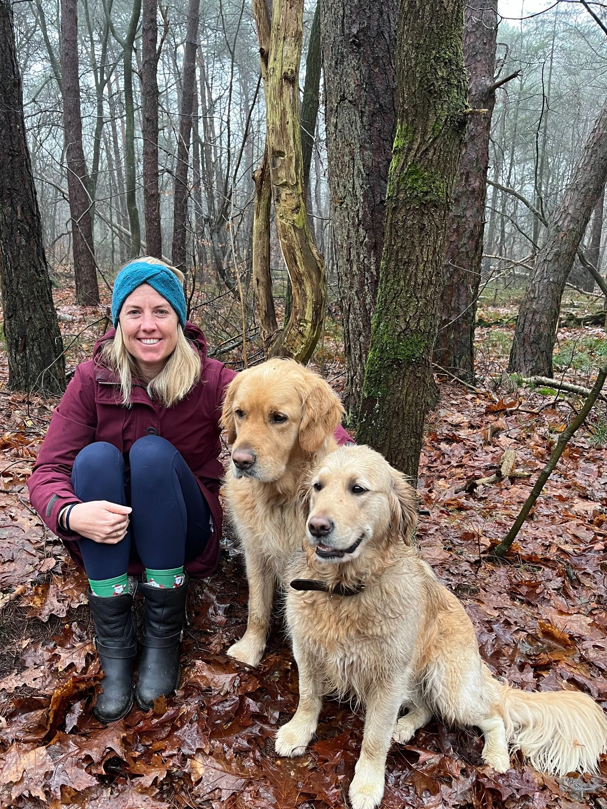 A woman in a maroon jacket and blue headband kneeling in a forest with two golden retrievers sitting next to her, surrounded by trees and wet autumn leaves.