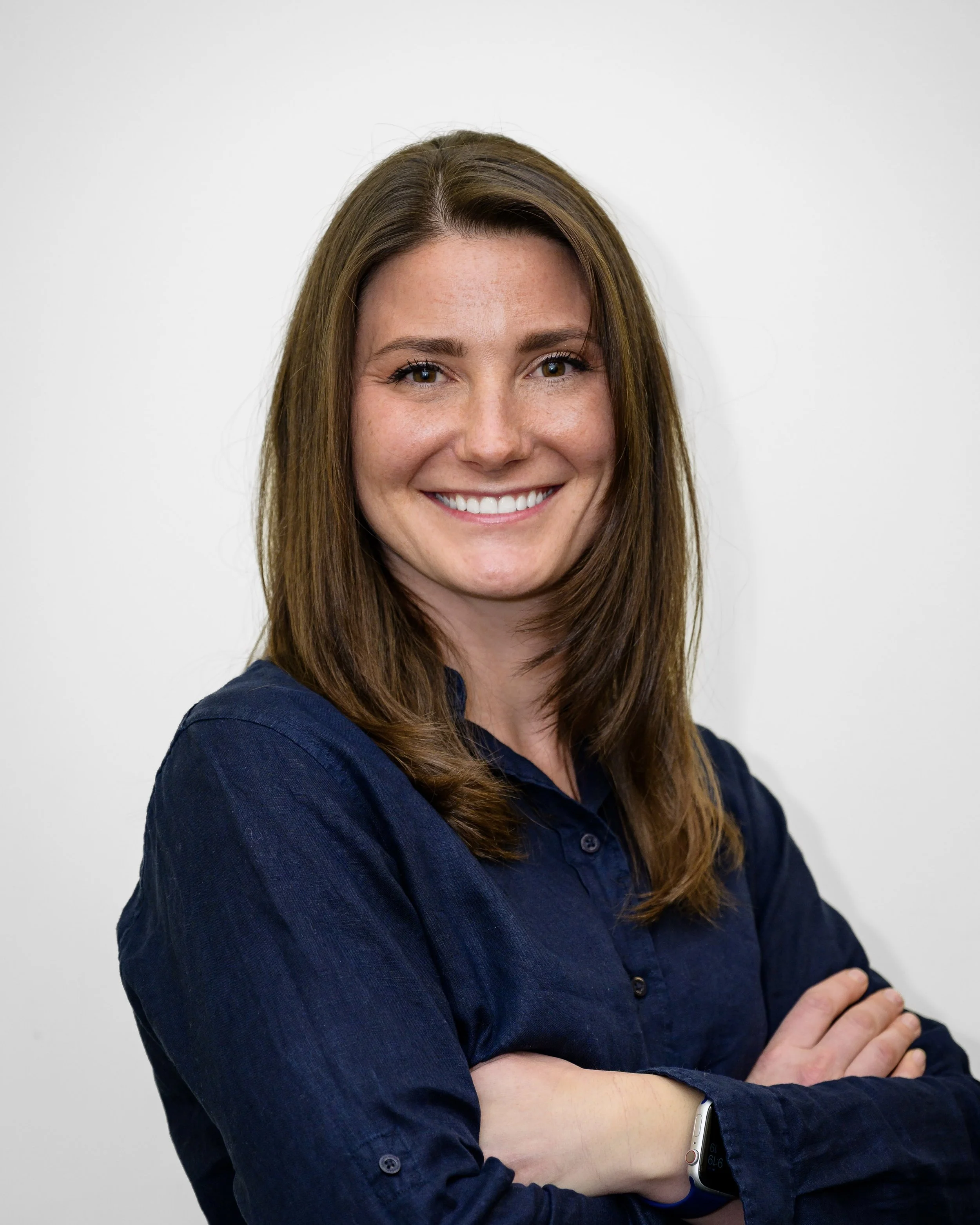 Portrait of a woman with long brown hair, smiling, wearing a dark blue shirt, standing against a plain white background.