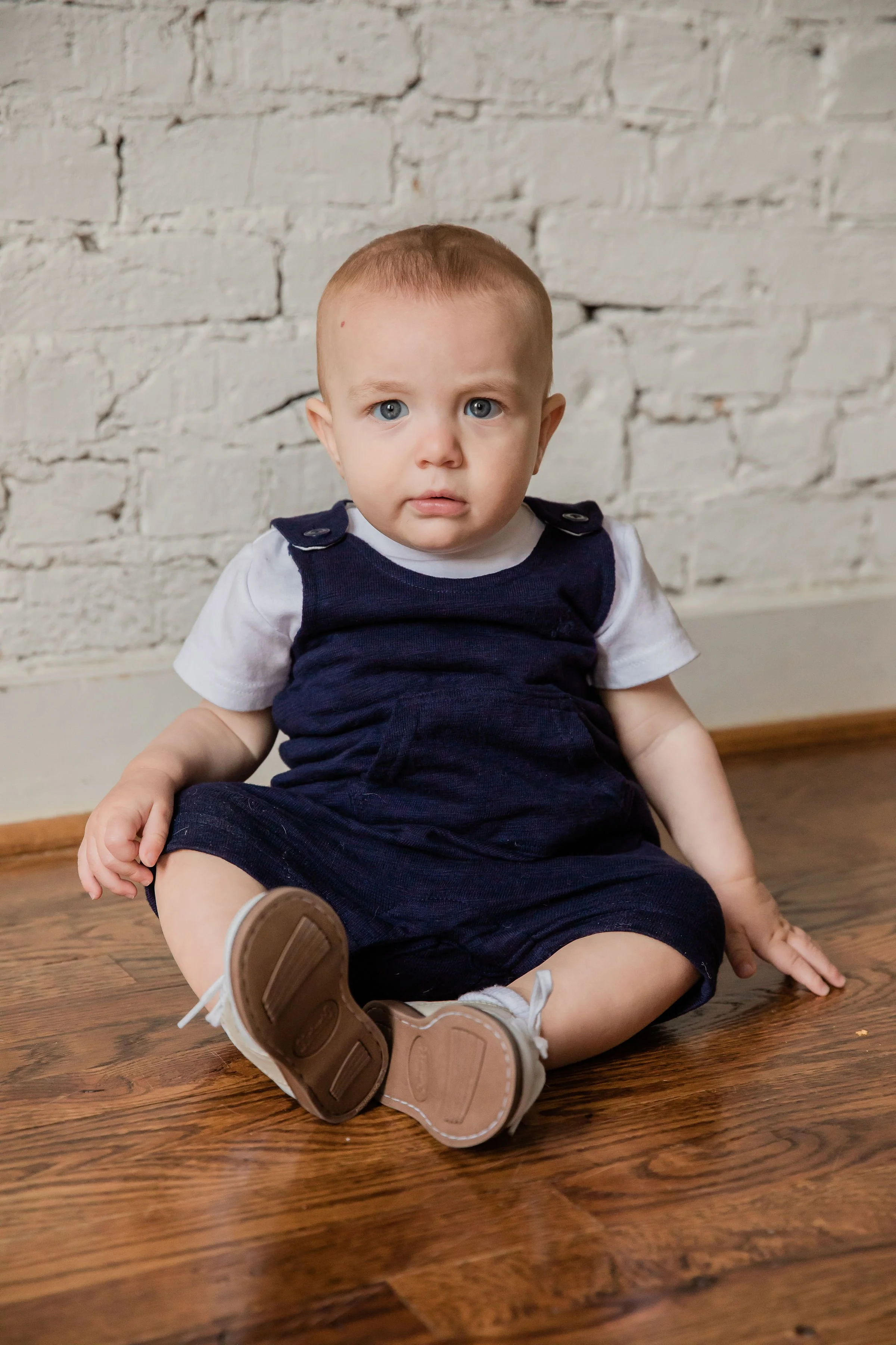 A young child with blue eyes and light brown hair sitting on a wooden floor, wearing a navy blue romper over a white shirt, and brown shoes, in front of a white brick wall.