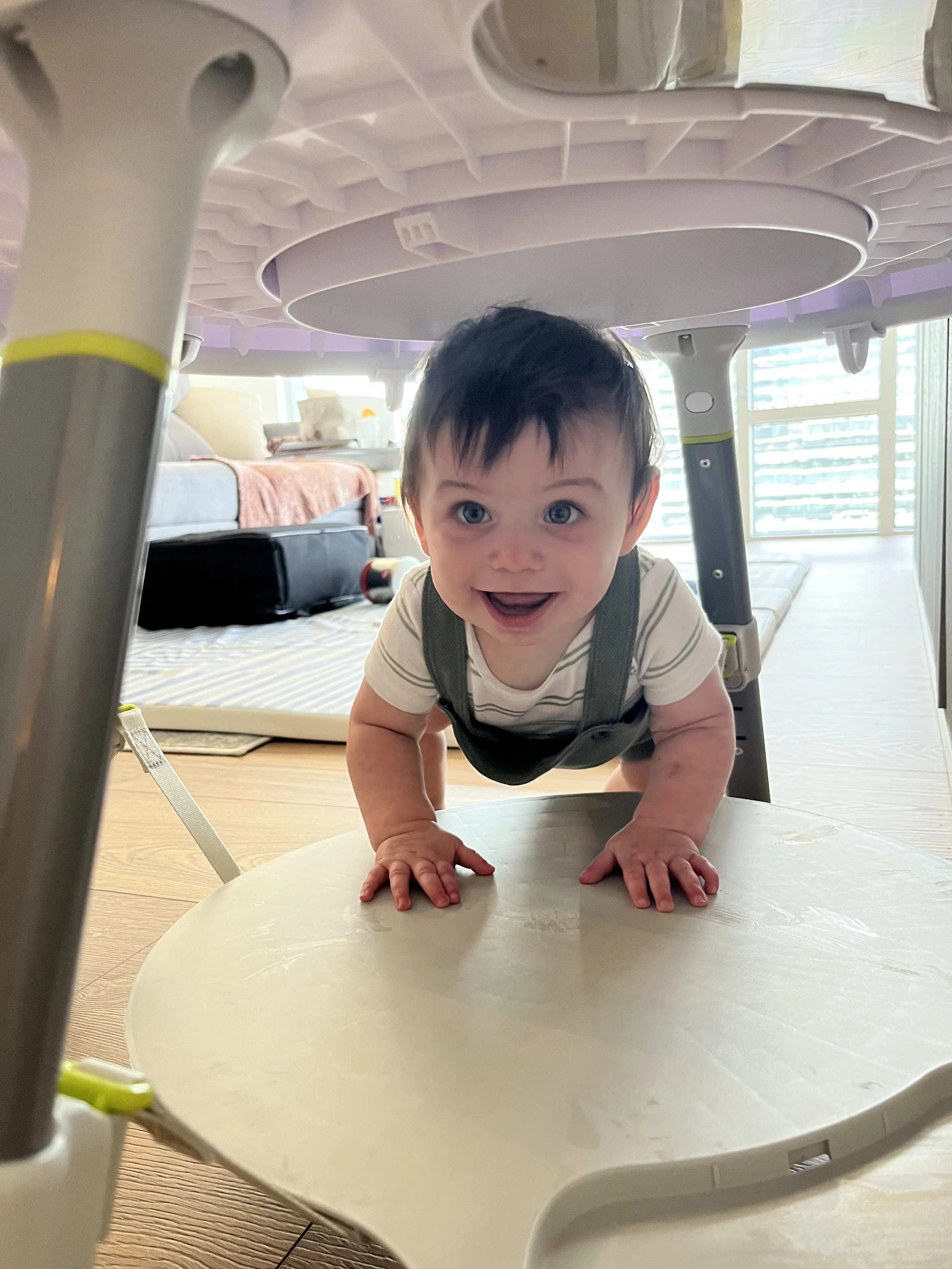 A smiling baby with dark hair and blue eyes crawling on a white table, viewed from underneath a child’s activity table.