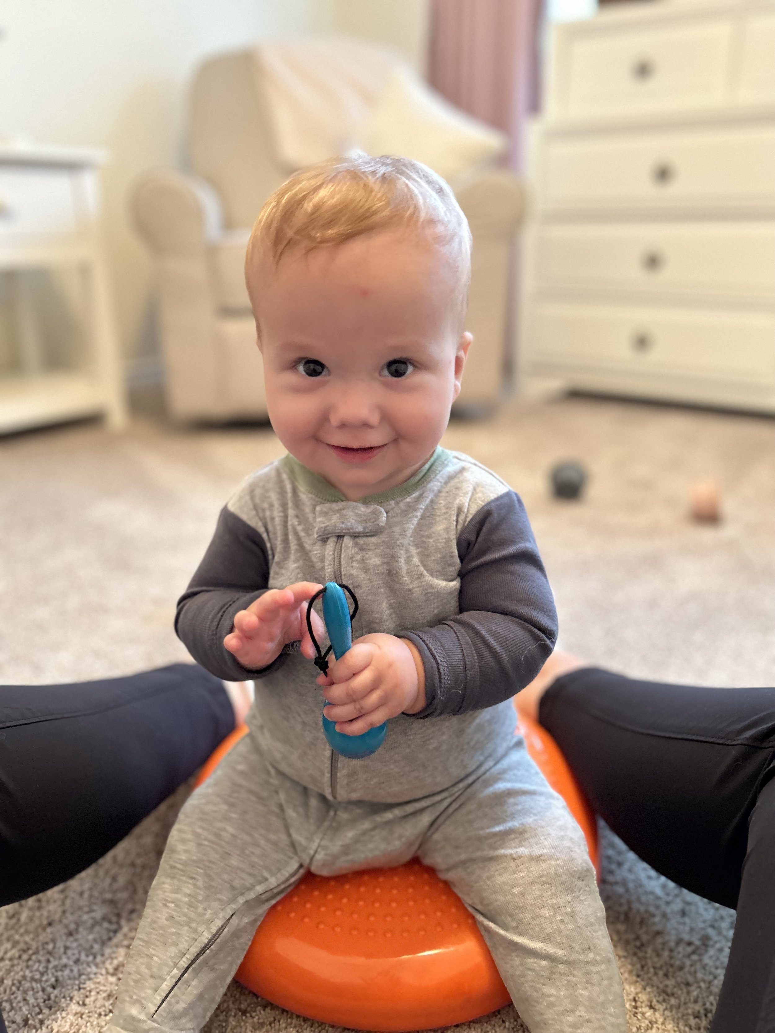 A young child with red hair, wearing a grey and dark grey pajama, sitting on an orange balance disc on a carpeted floor, smiling and holding a blue toy.