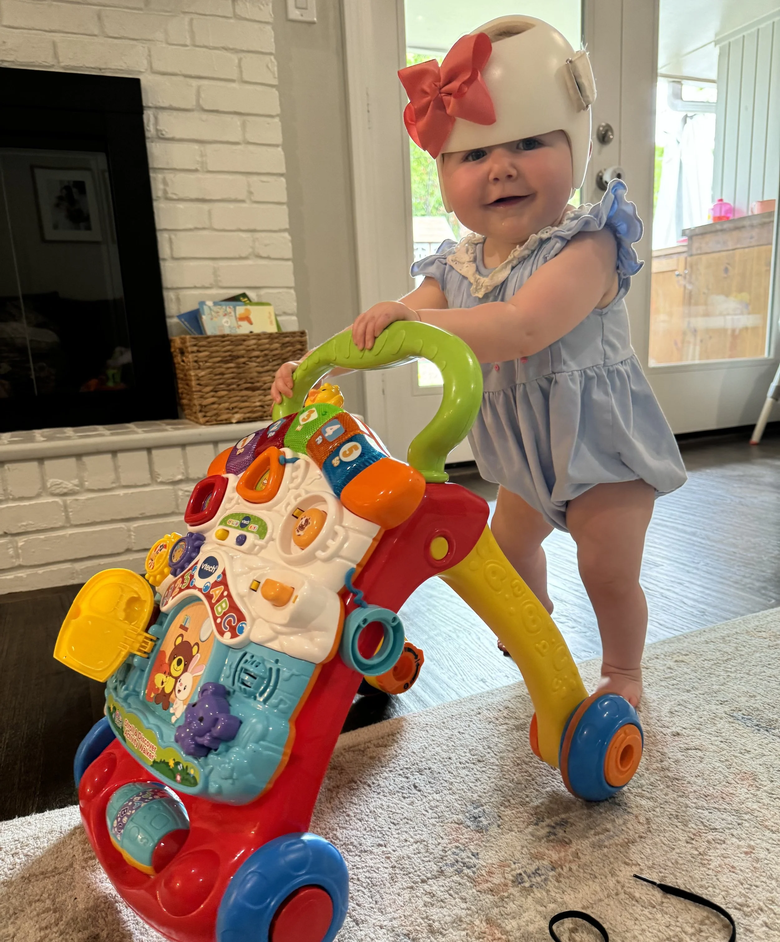A smiling baby girl standing behind a colorful plastic push toy with various buttons and shapes, wearing a light blue ruffled dress and a white helmet with a big red bow, inside a home near a door with a window.