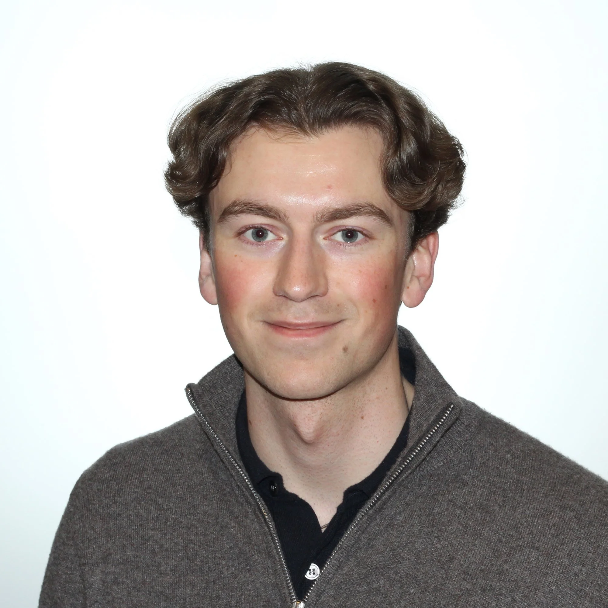 Henkilö looking suoraan kameraan, hymyilevä, ruskea neule ja musta paita, valoisa tausta.