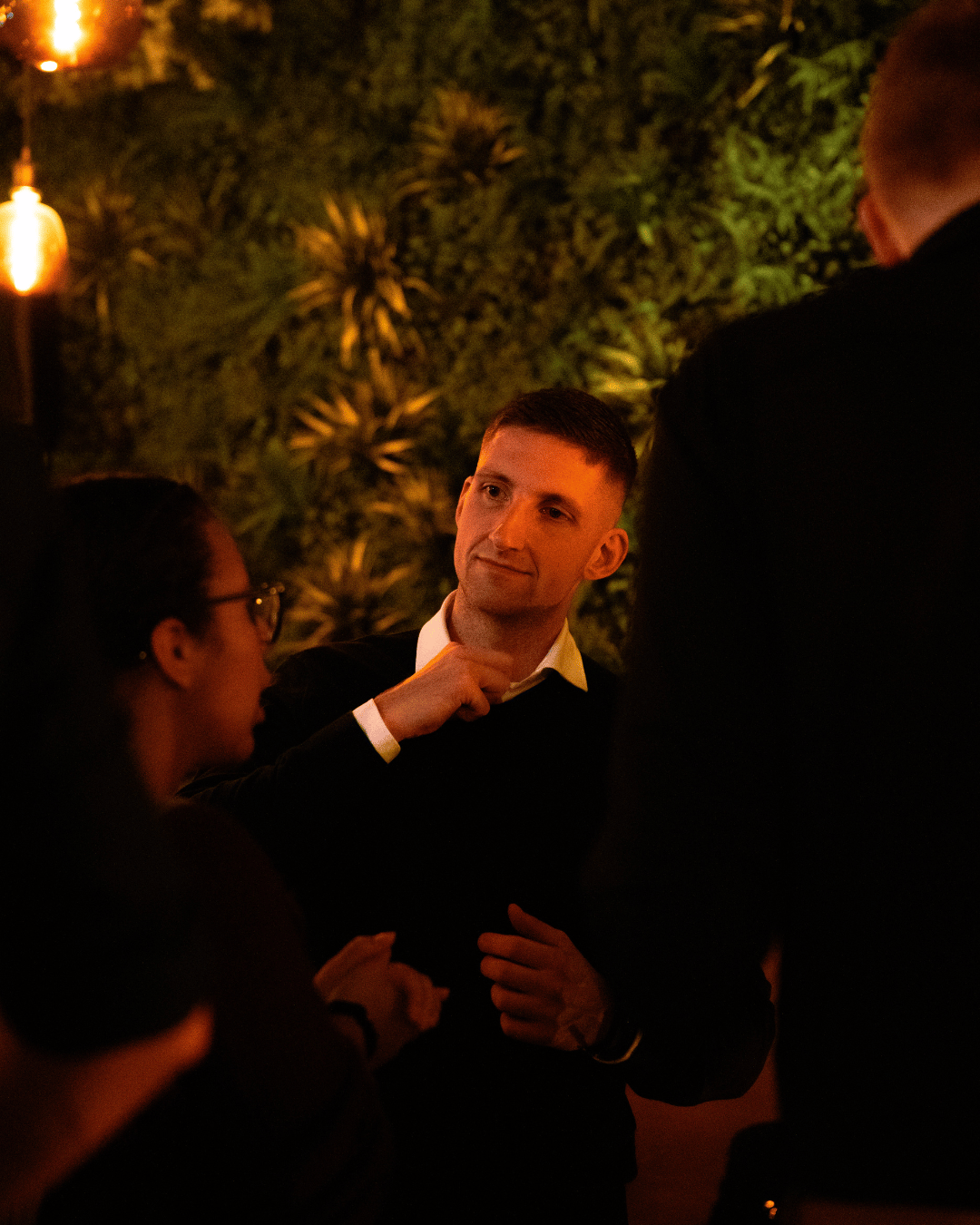 Drie mensen hebben een gesprek in een dimverlichte ruimte met groene muur en warme verlichting.