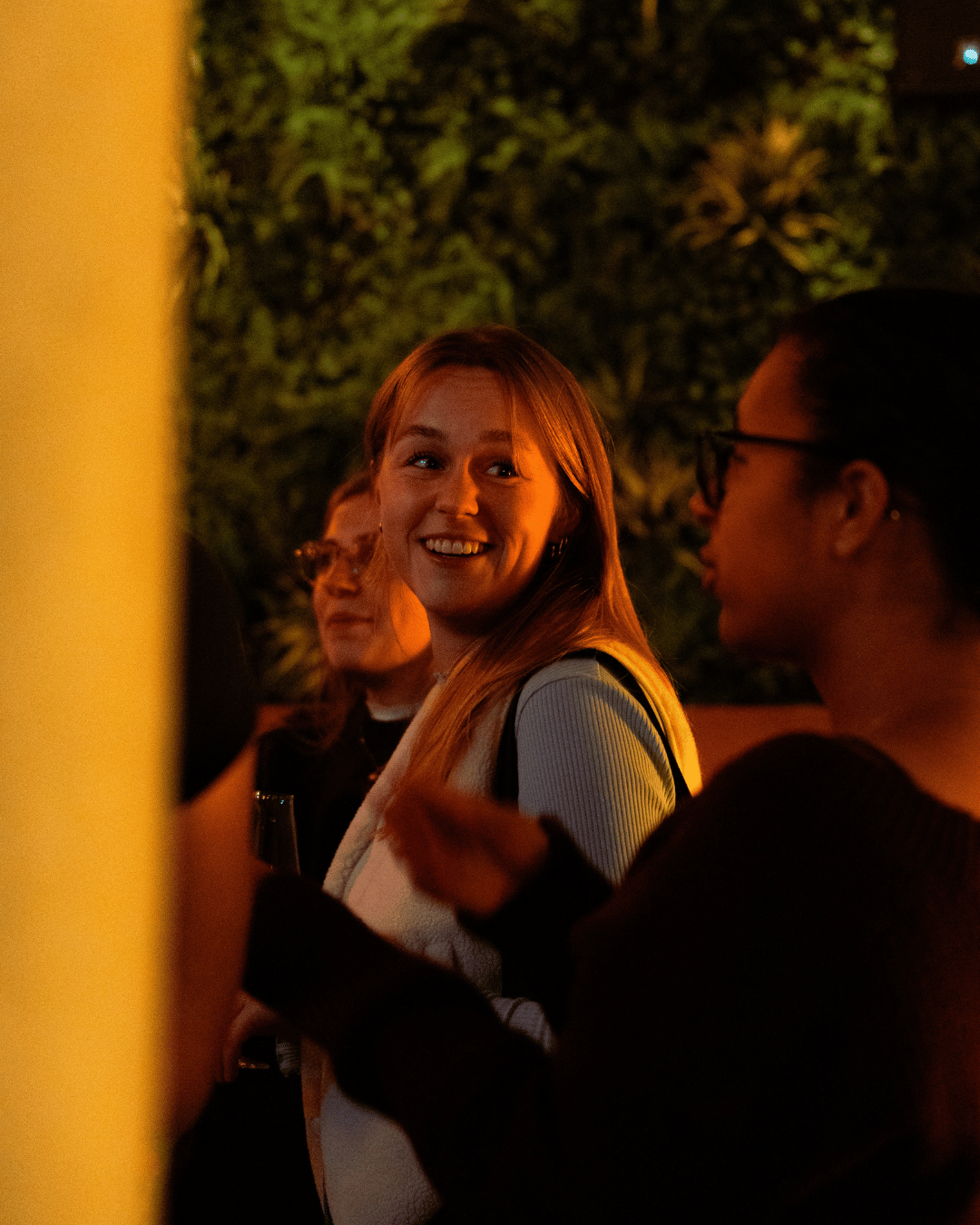 Vrouw lacht en praat met anderen tijdens een avond in een tuin of patio met groene planten op de achtergrond.