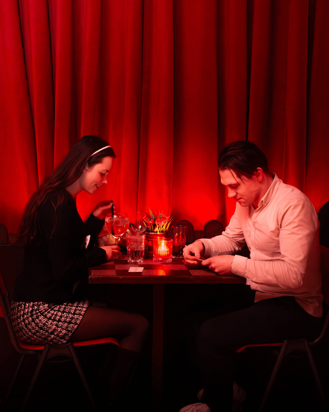 Een man en vrouw zitten tegenover elkaar aan een tafel in een restaurant met zacht roodverlicht decor. Ze lijken te praten en hebben drinken voor zich.