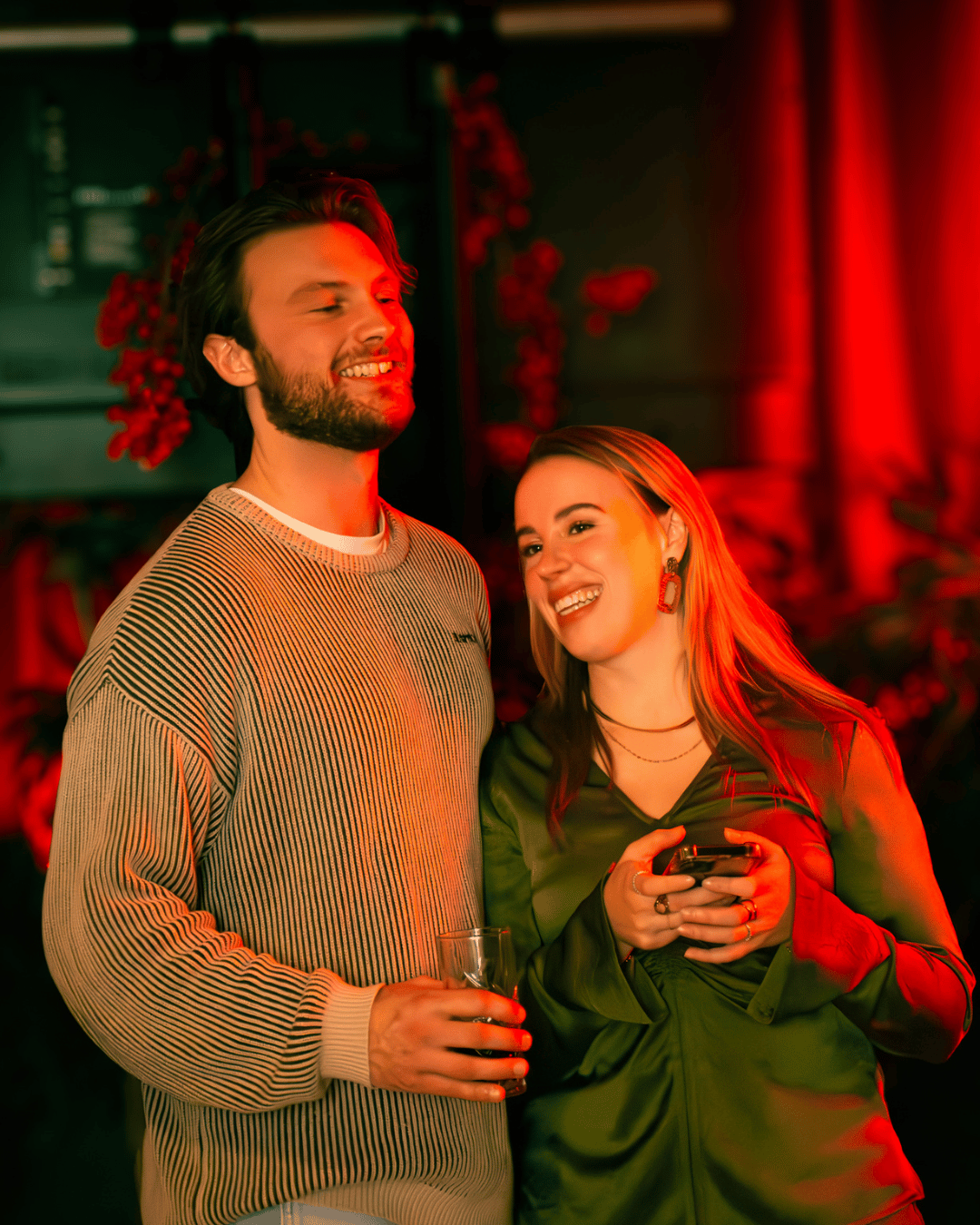 Twee mensen lachen en praten samen tijdens een feestje, een man met een glas in de hand en een vrouw met een smartphone, met felrode verlichting op de achtergrond.