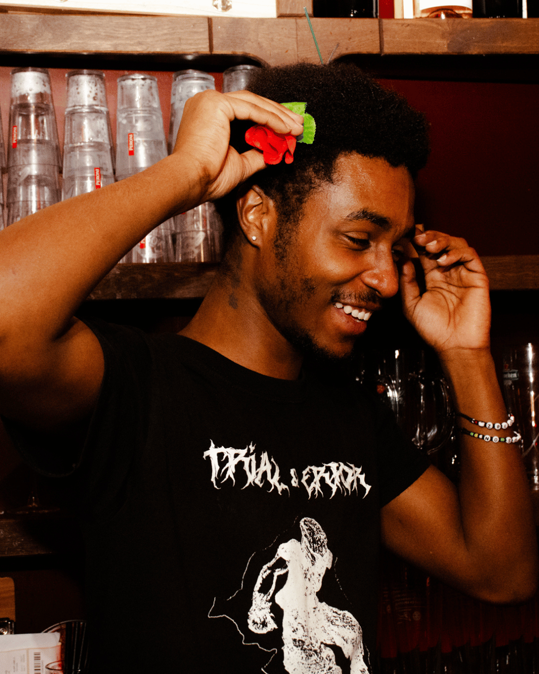 Man met een zwarte T-shirt met een grafisch ontwerp, lachend, met een haarborstel in de hand, in een bar of café.
