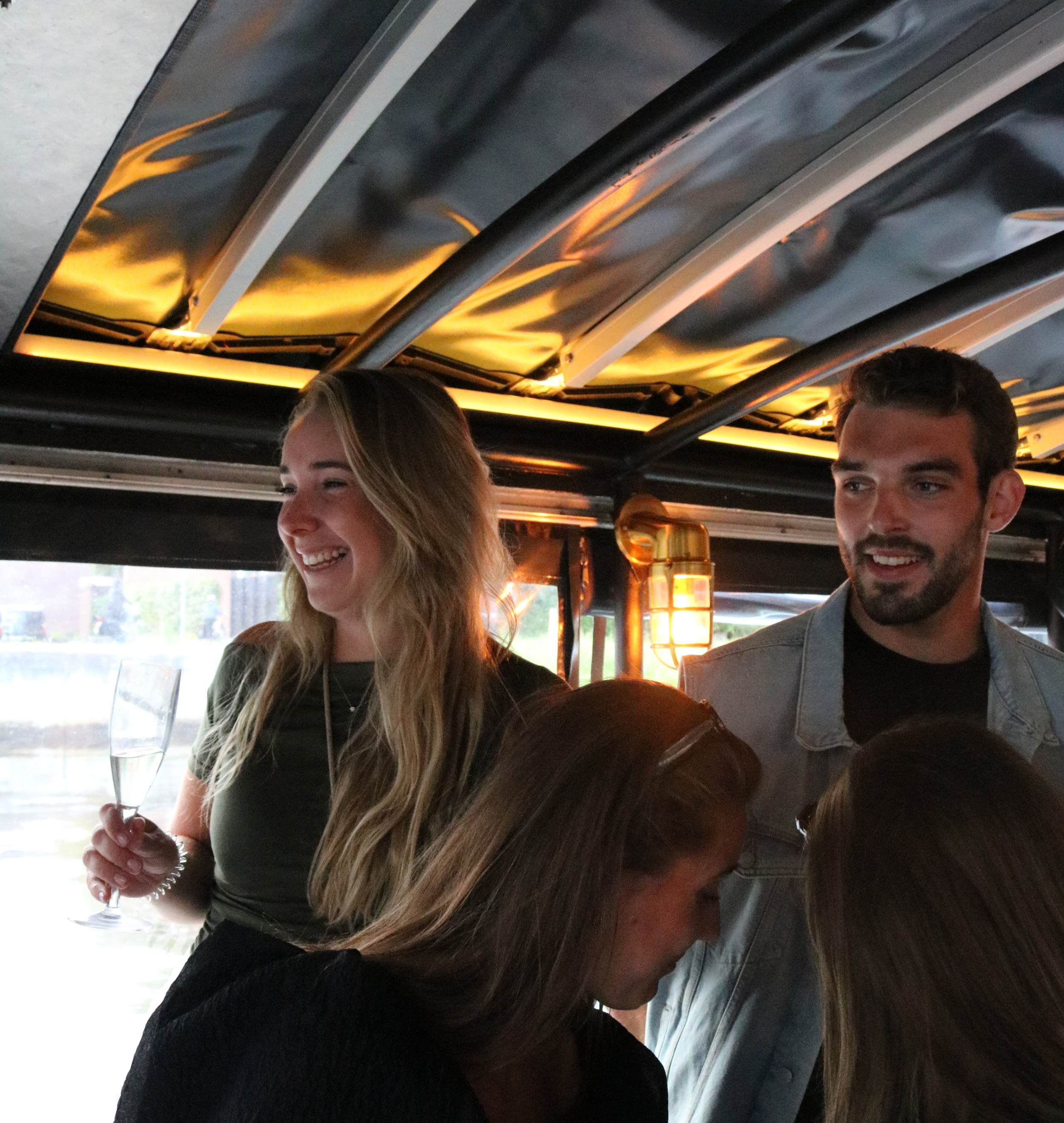 Verrassende groepsfoto van vier mensen in een boot ingericht met een industrieel plafond en warme verlichting. Een blonde vrouw anderhalf glimlachend, houdt een champagneglas en staat naast een donkere vrouw. Een man met getinte huid en een baard kijkt naar de vrouwen. Twee andere mensen zitten met hun hoofden naar beneden, niet volledig zichtbaar.
