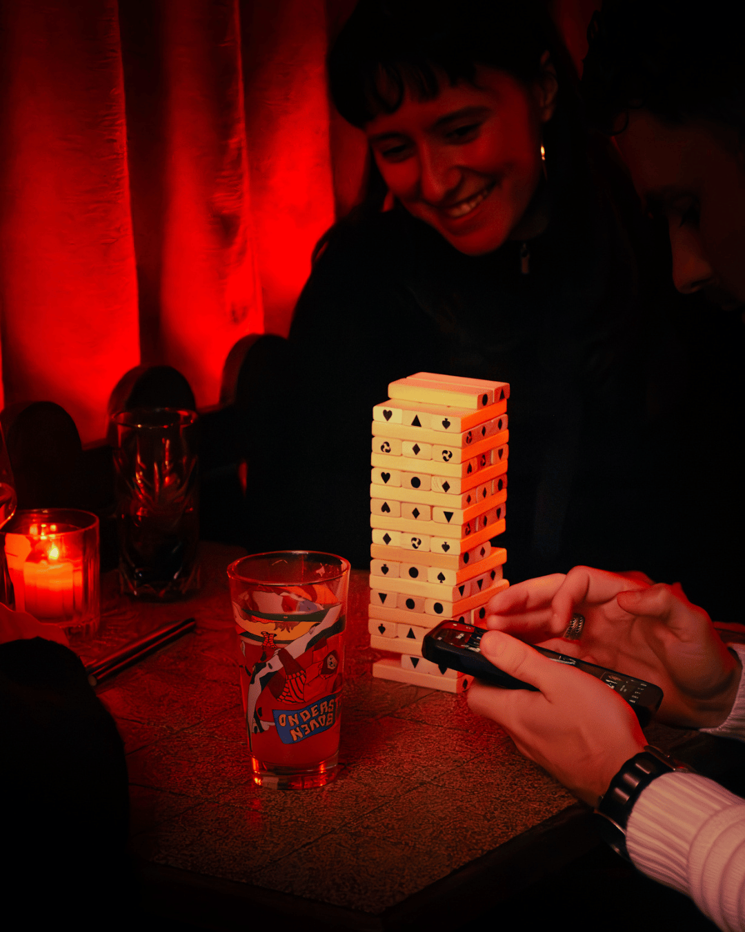 Twee mensen spelen een spel met een toren van dominostenen op een tafel, verlicht door rood licht, terwijl iemand een apparaat vasthoudt.