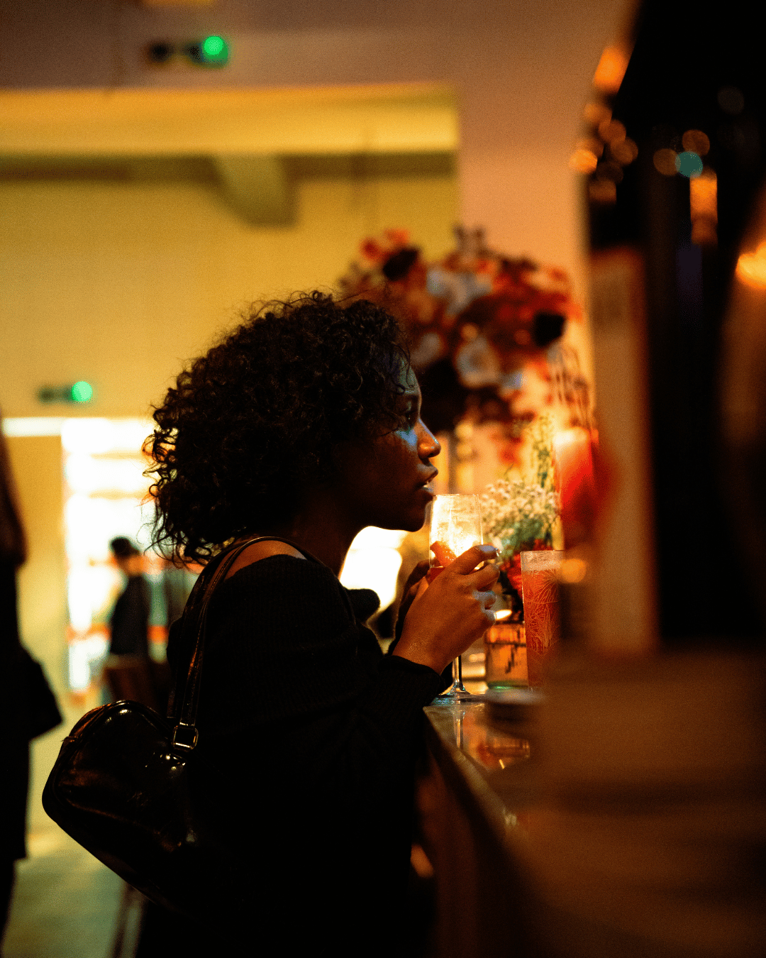 Een vrouw met krullend haar drinkt uit een wijnglas in een bar met warme verlichting, op de achtergrond mensen en decoraties.