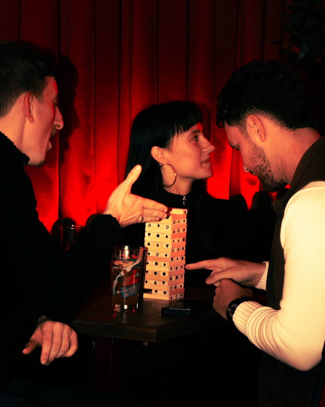 Drie mensen die binnen een bar of club spelen een spelletje Jenga op een tafel, met een glas drinken erbij, tegen een achtergrond van rood gordijn.