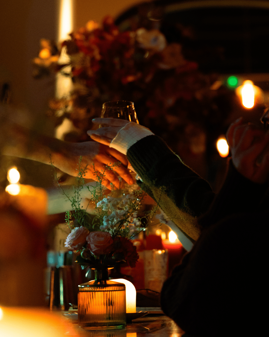 Mensen die een toast uitwisselen aan een sfeervol gedekte tafel met kaarslicht en bloemen, binnen met zachte verlichting.