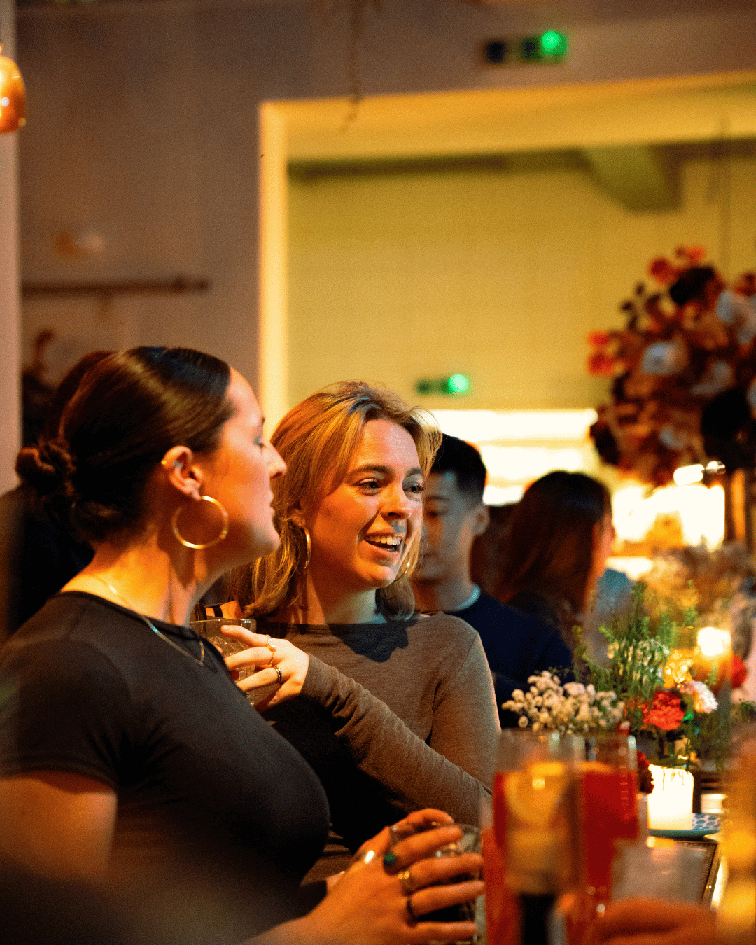 Mensen genieten samen aan de bar in een sfeervol restaurant, omgeven door bloemen en kaarslichten.