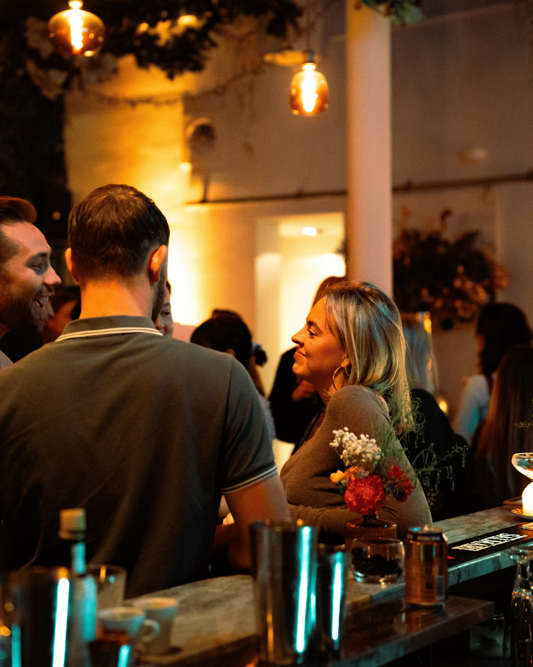Mensen die in een bar of restaurant praten en lachen, met warme sfeerverlichting en bloemen op de tafel.