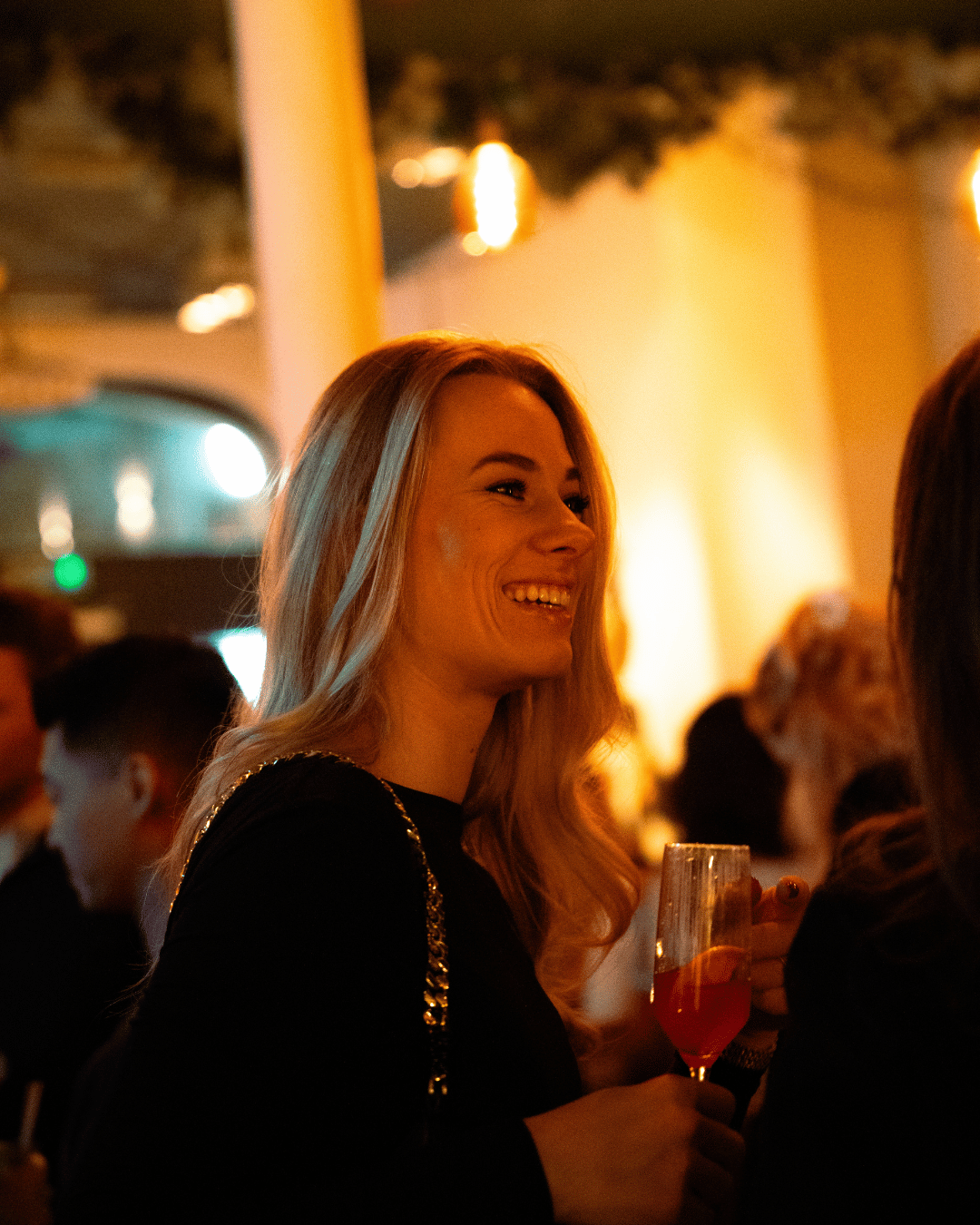 Een lachende vrouw met lang blond haar houdt een glas met een roze drankje in een gezellige, warme sfeer. Ze is gekleed in een zwarte outfit met metallic details.