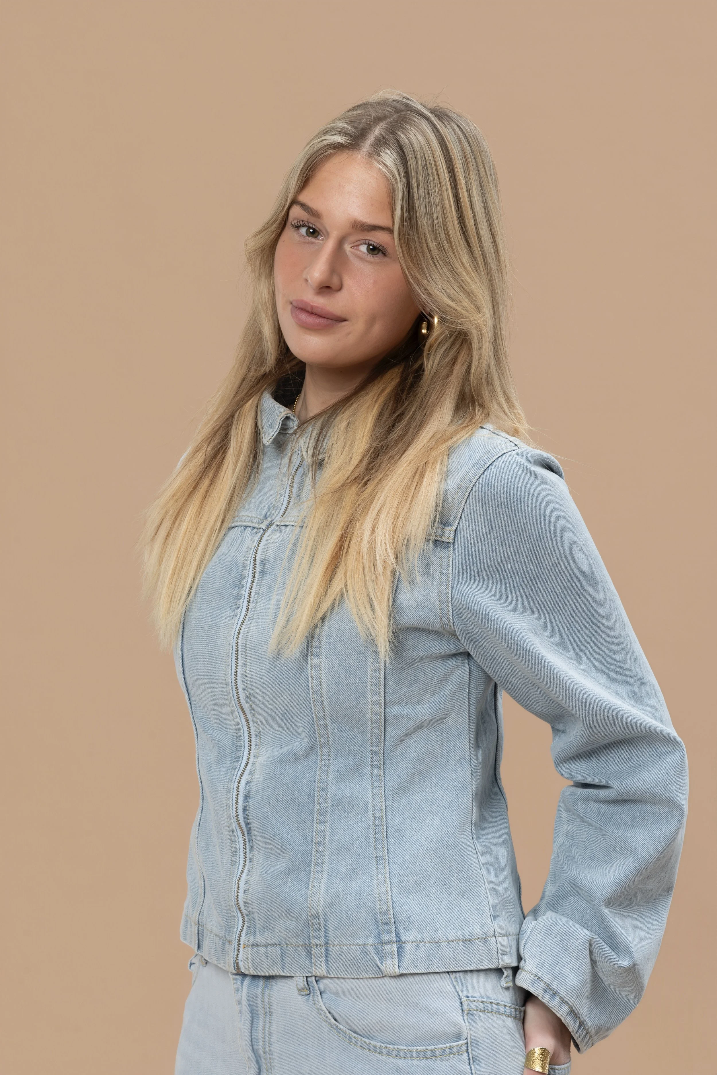 Portret van een jonge vrouw met lang blond haar, in een licht blauwe denim jas en spijkerbroek, poseert tegen een beige achtergrond.