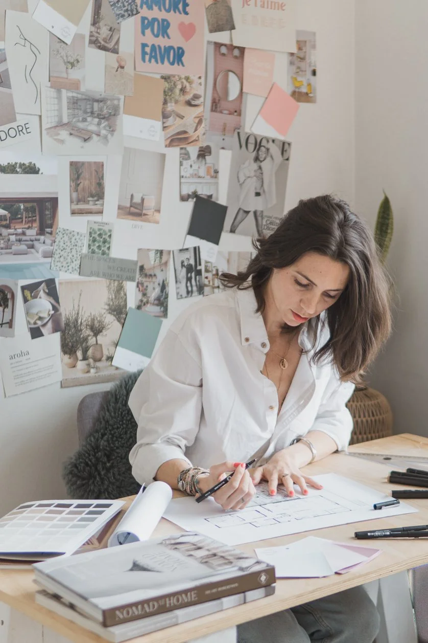 Woman sitting at a wooden desk working on design sketches with color palettes and design magazines, and a wall behind her decorated with photos, mood boards, and magazine clippings.